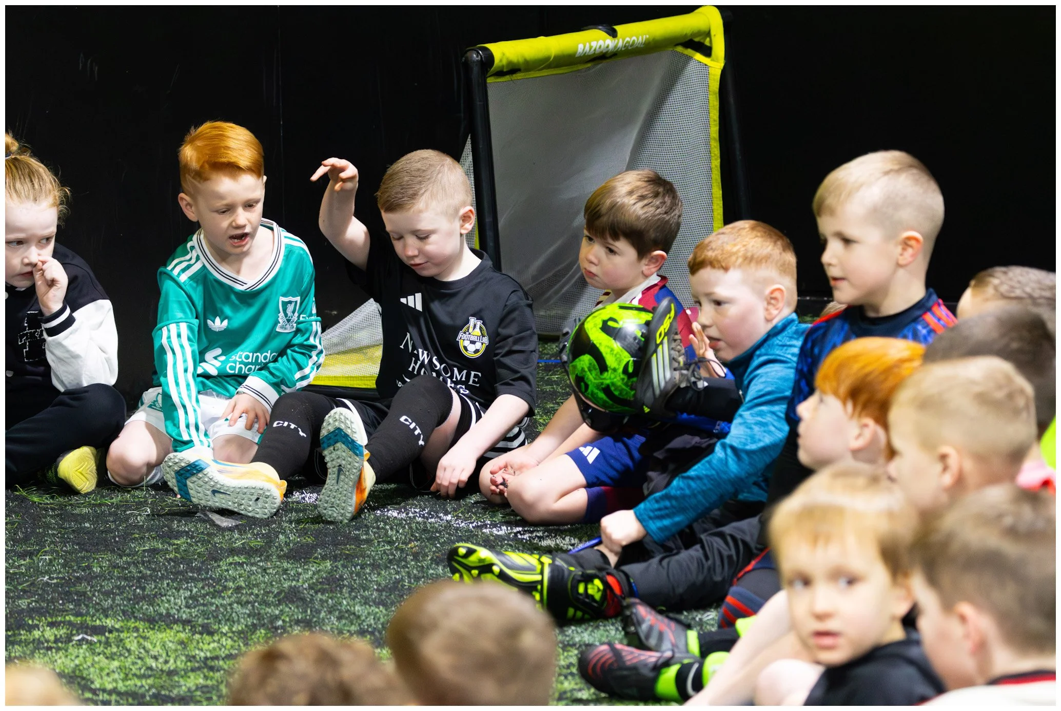 Group of young children sitting on indoor turf, listening to a girl in sporty outfit with blonde hair, as she speaks with her hand raised.