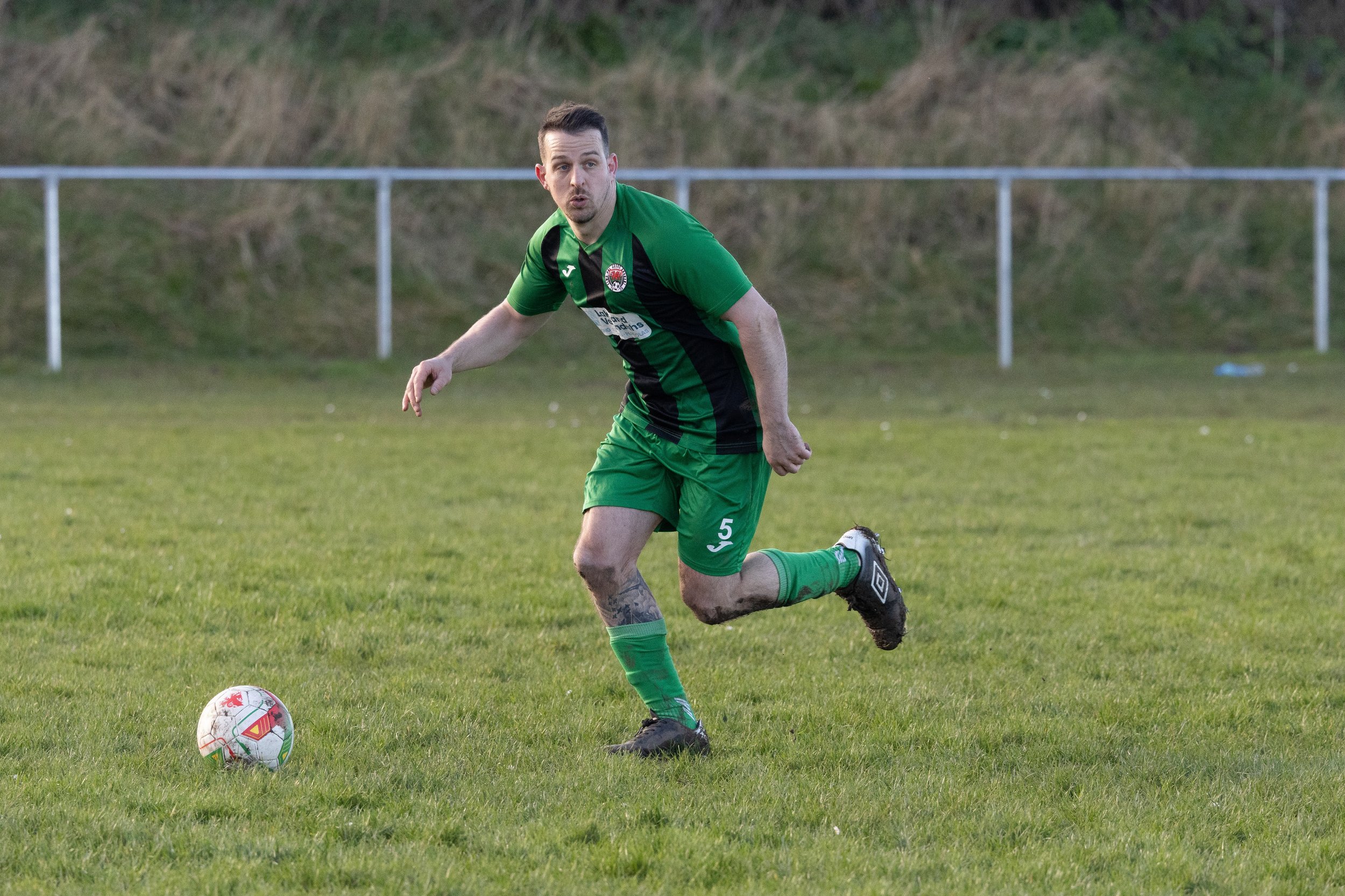 A football player in green and black uniform is on the field, running towards a soccer ball.