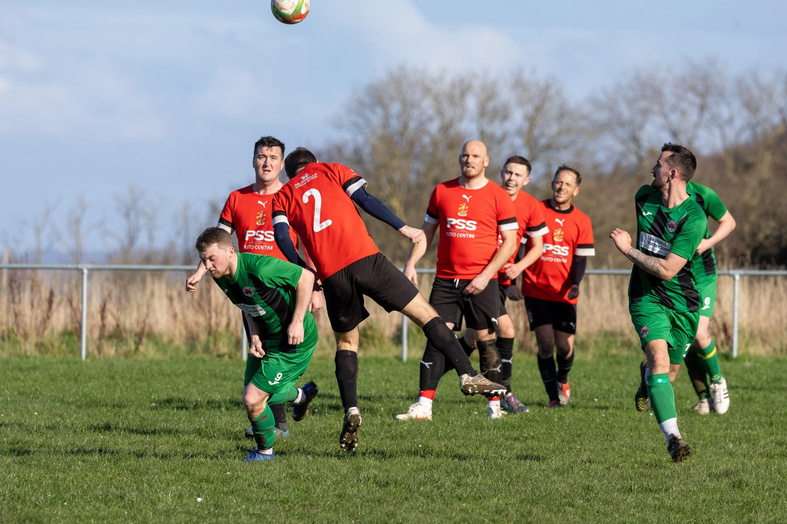 Soccer players in red and green jerseys competing for the ball on a grassy field under a partly cloudy sky.