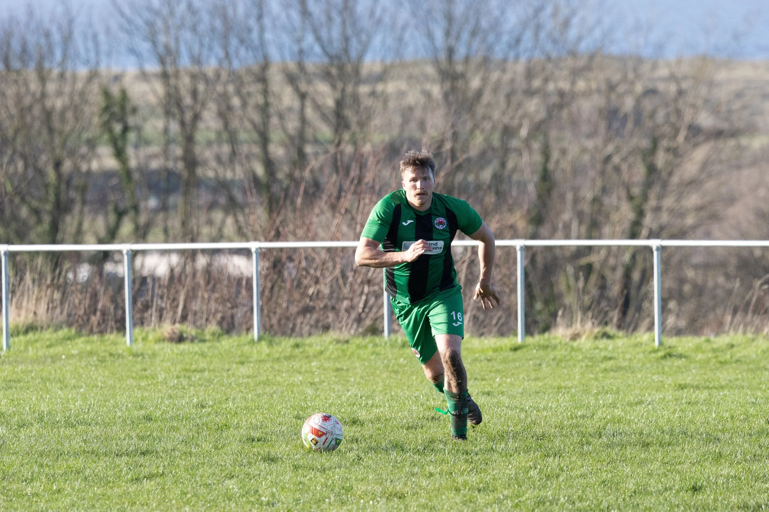 A soccer player in green jersey running on a grass field with a soccer ball at his feet, behind a white fence and leafless trees.