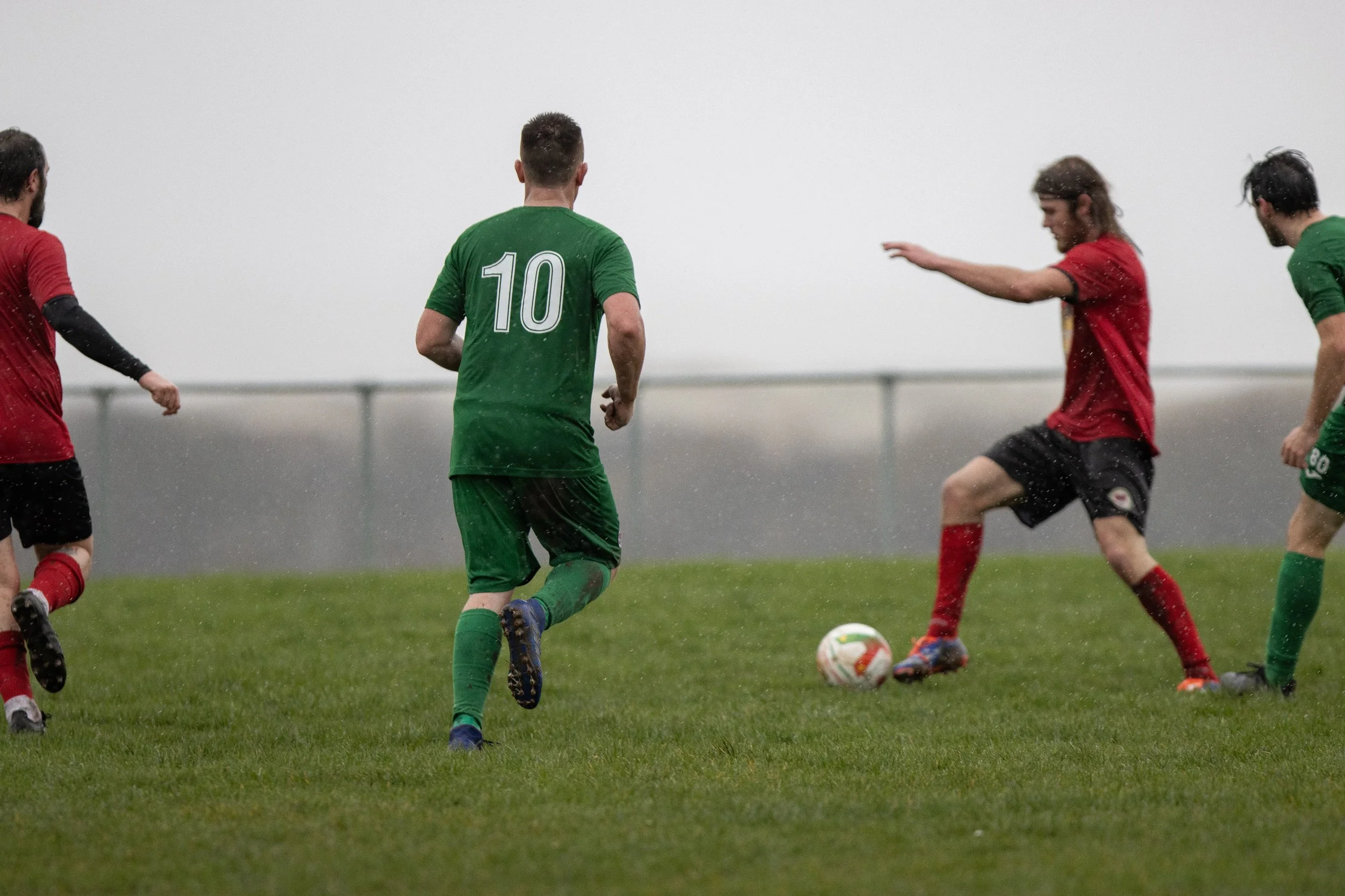 Soccer players in red and green uniforms playing in the rain on a grassy field, competing for the ball.