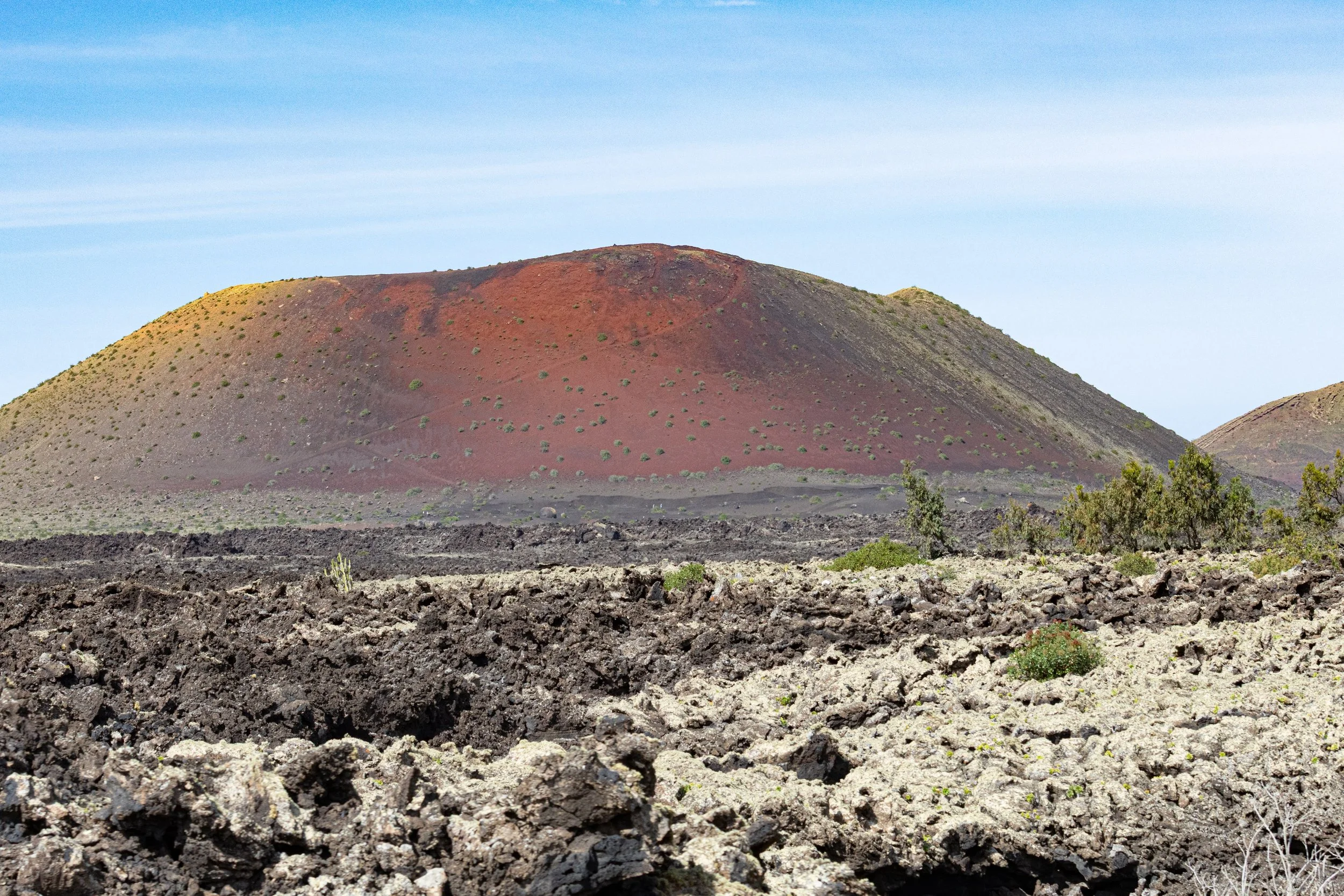 A volcanic landscape with a large, colorful volcano in the background and rough, dark lava rocks in the foreground, with sparse vegetation and a clear blue sky.