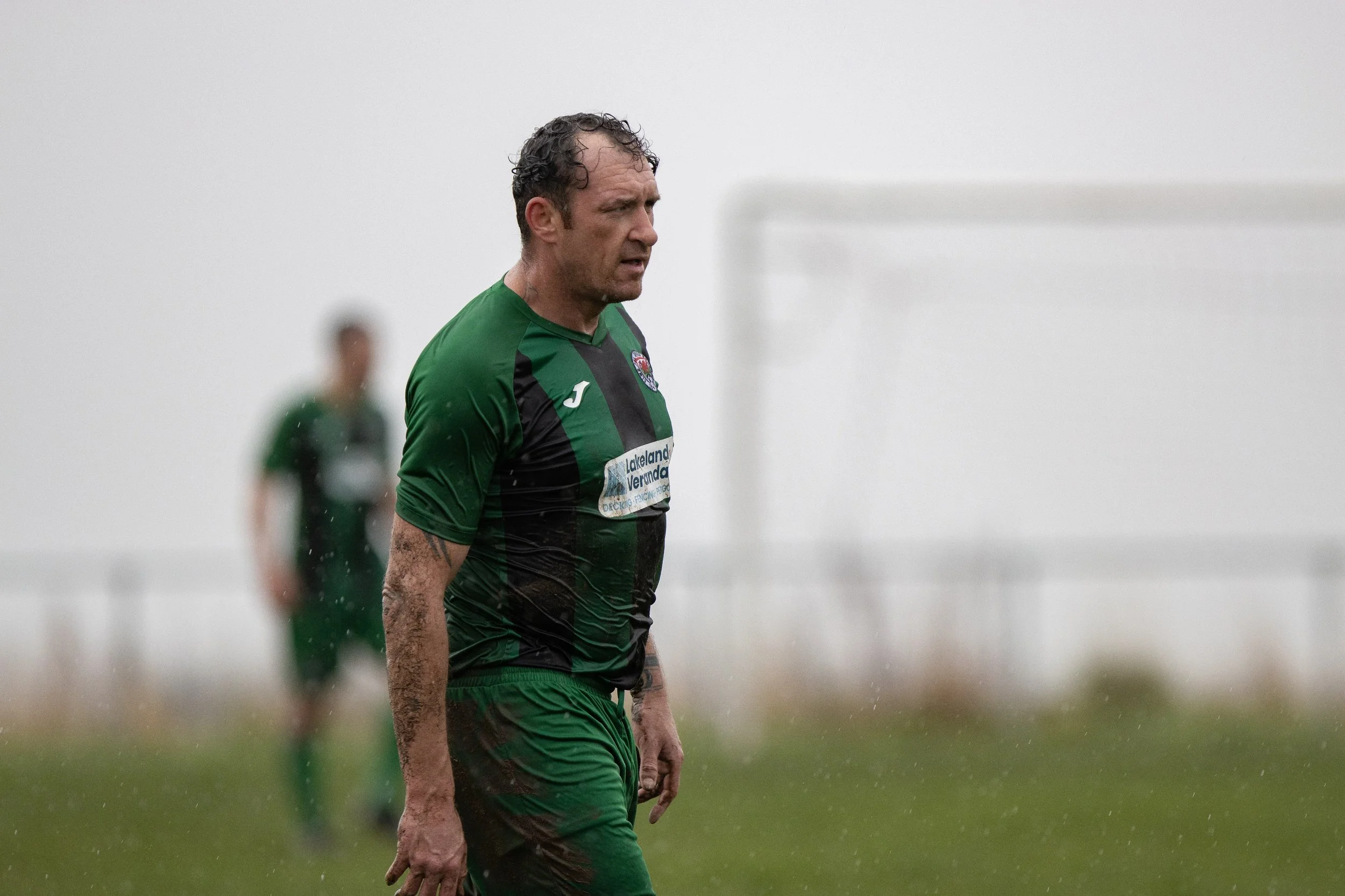 A male soccer player in a green and black uniform walking on a muddy field with a foggy background and blurred teammate in the distance.