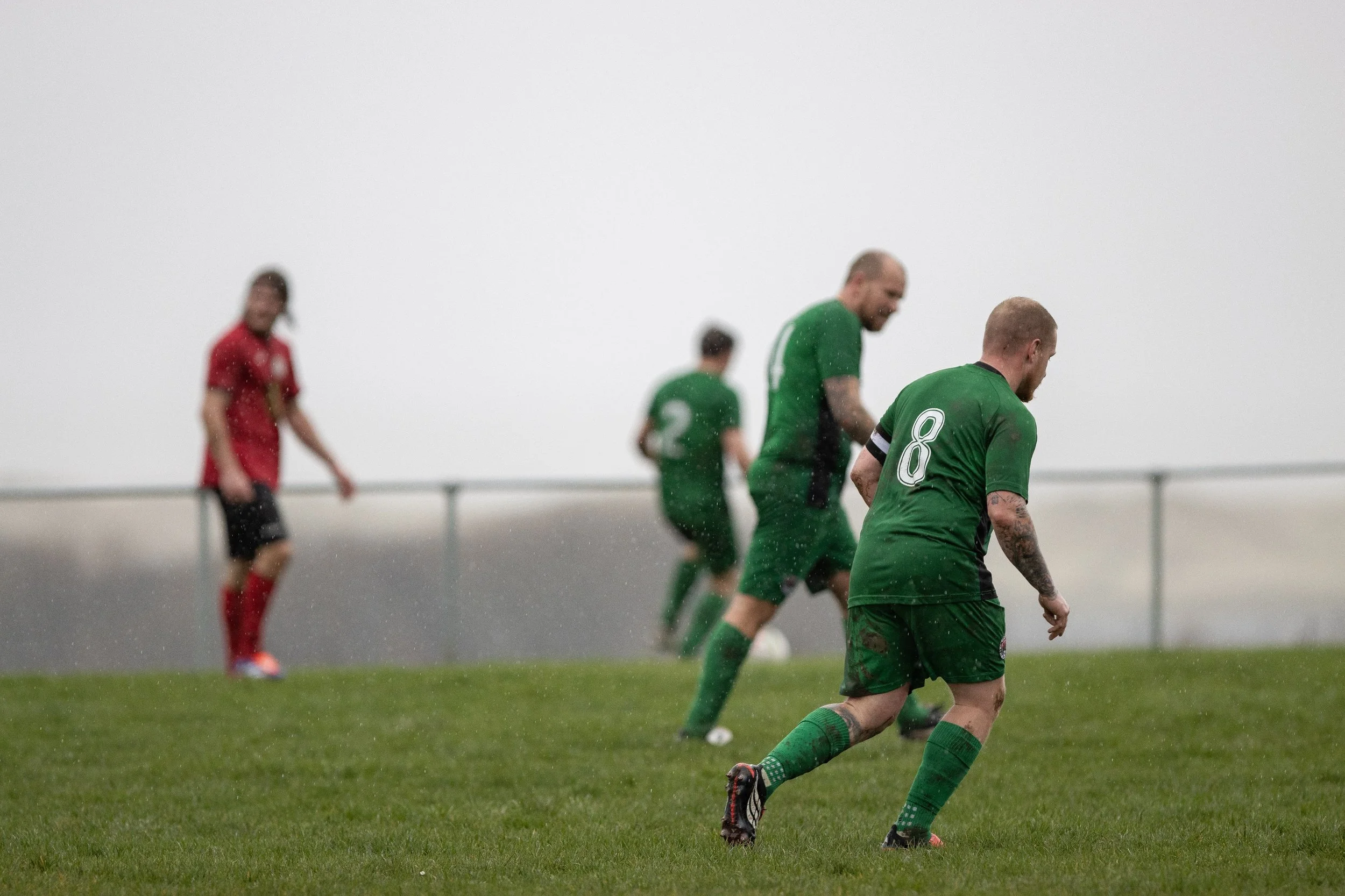 Soccer players on a rainy field, with one in the foreground wearing a green jersey with the number 8, and others in the background, some in red jerseys, under gray, foggy skies.