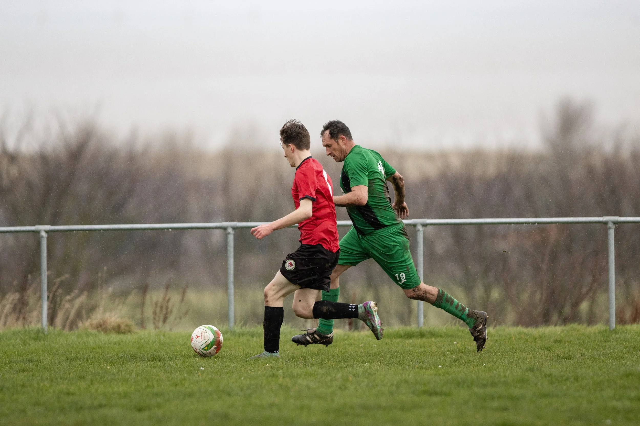 Two soccer players compete for the ball on a grassy field, with one wearing a red jersey and black shorts, and the other in a green jersey and shorts. There is a metal fence in the background amidst a cloudy, overcast sky.