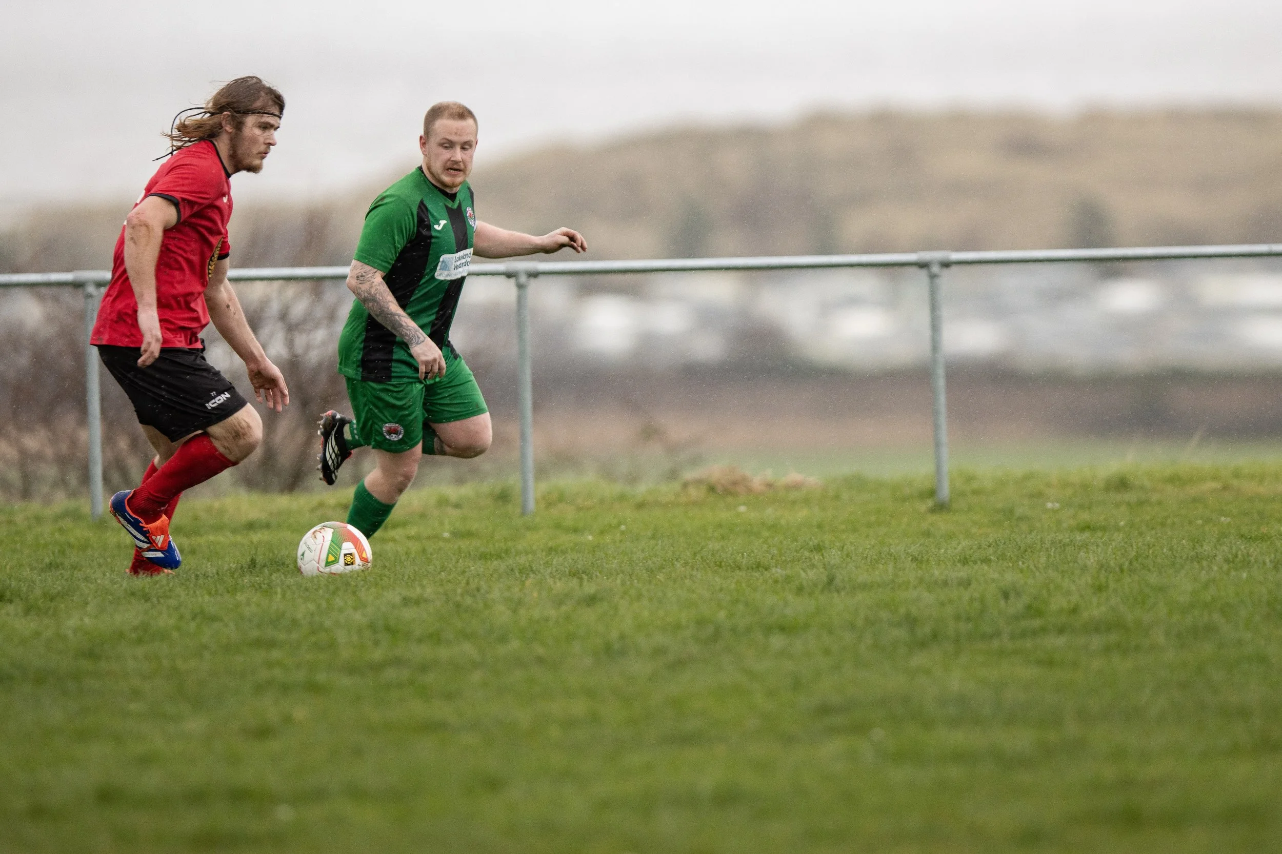 Two soccer players on a grassy field chasing a soccer ball, with a metal fence and overcast sky in the background.