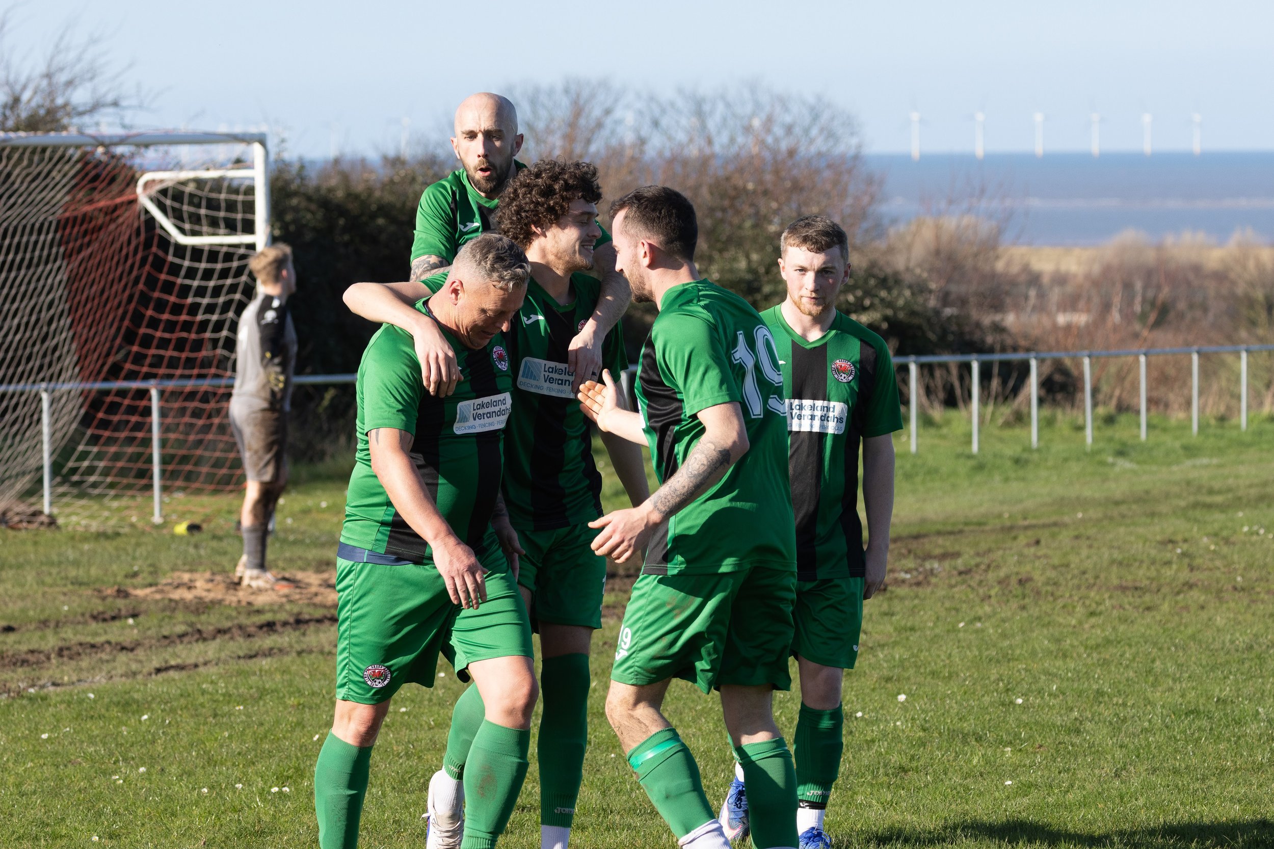 A group of five male soccer players in green jerseys celebrating on the field after a goal, with a soccer goal and cloudy sky in the background.