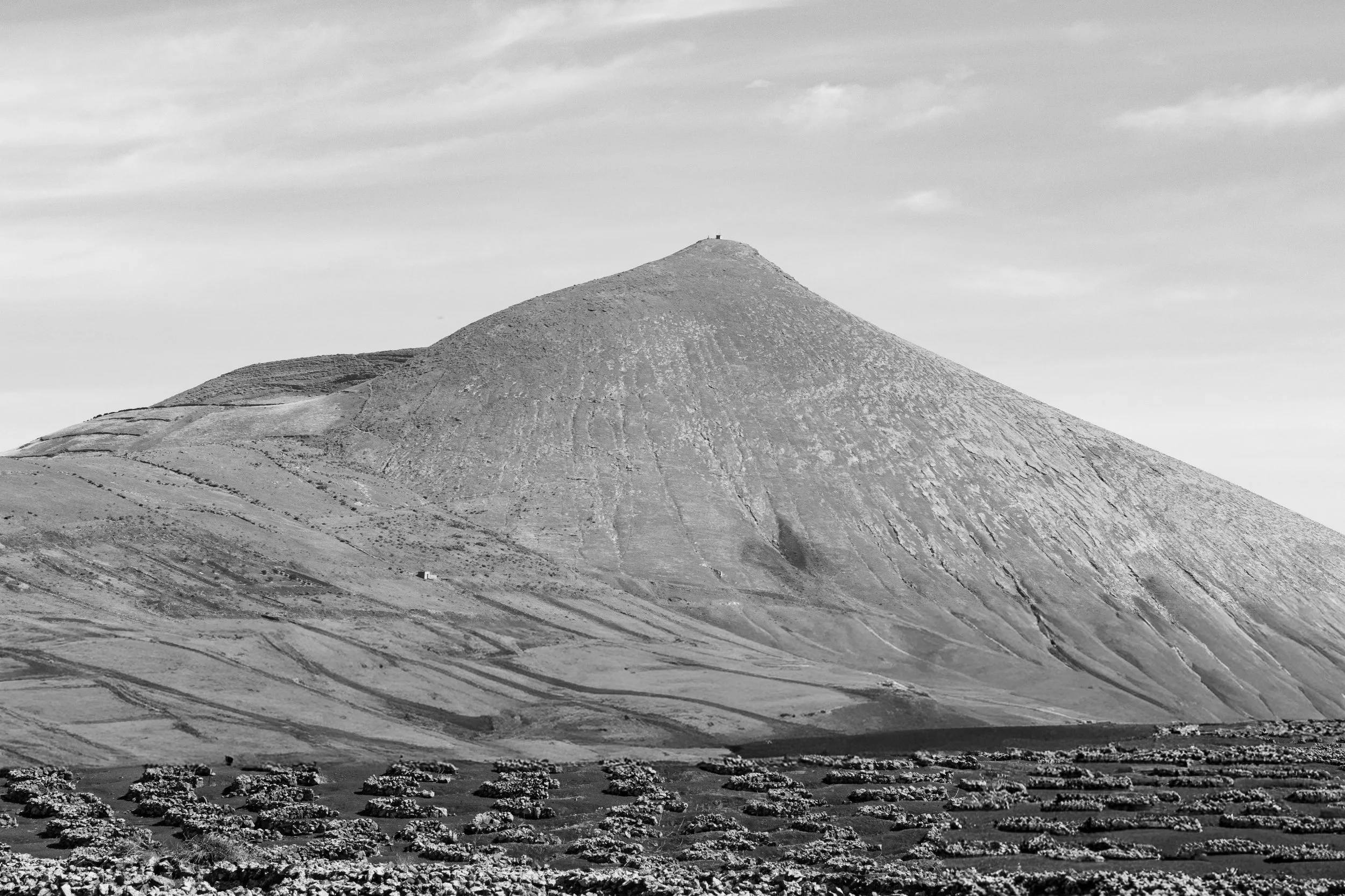 Black and white photo of a mountain with terraced fields at the base and a small structure near the top.
