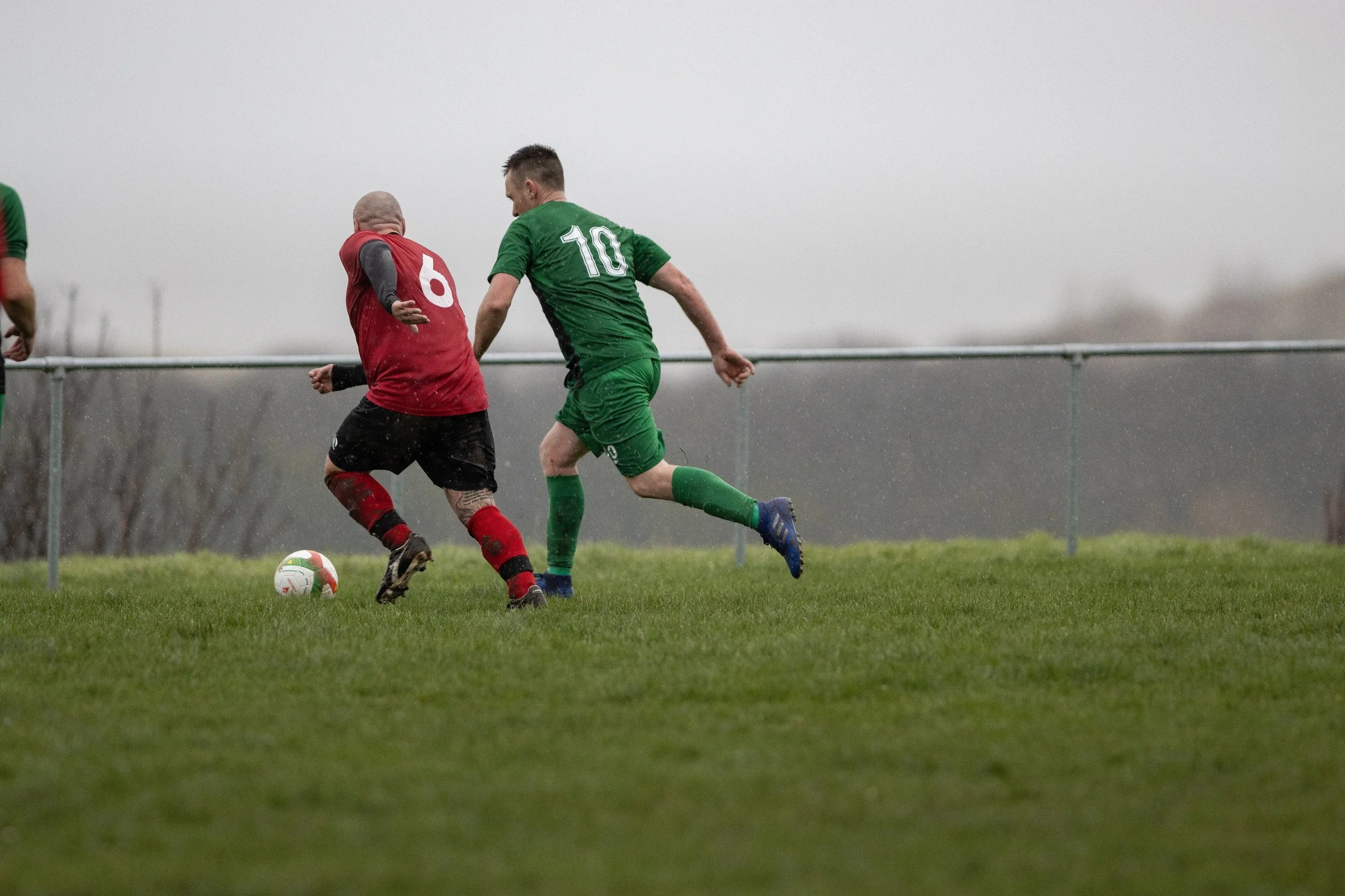 Two soccer players, one in a red jersey and the other in a green jersey, compete for the ball on a grassy field in rainy weather, with a fence and cloudy sky in the background.