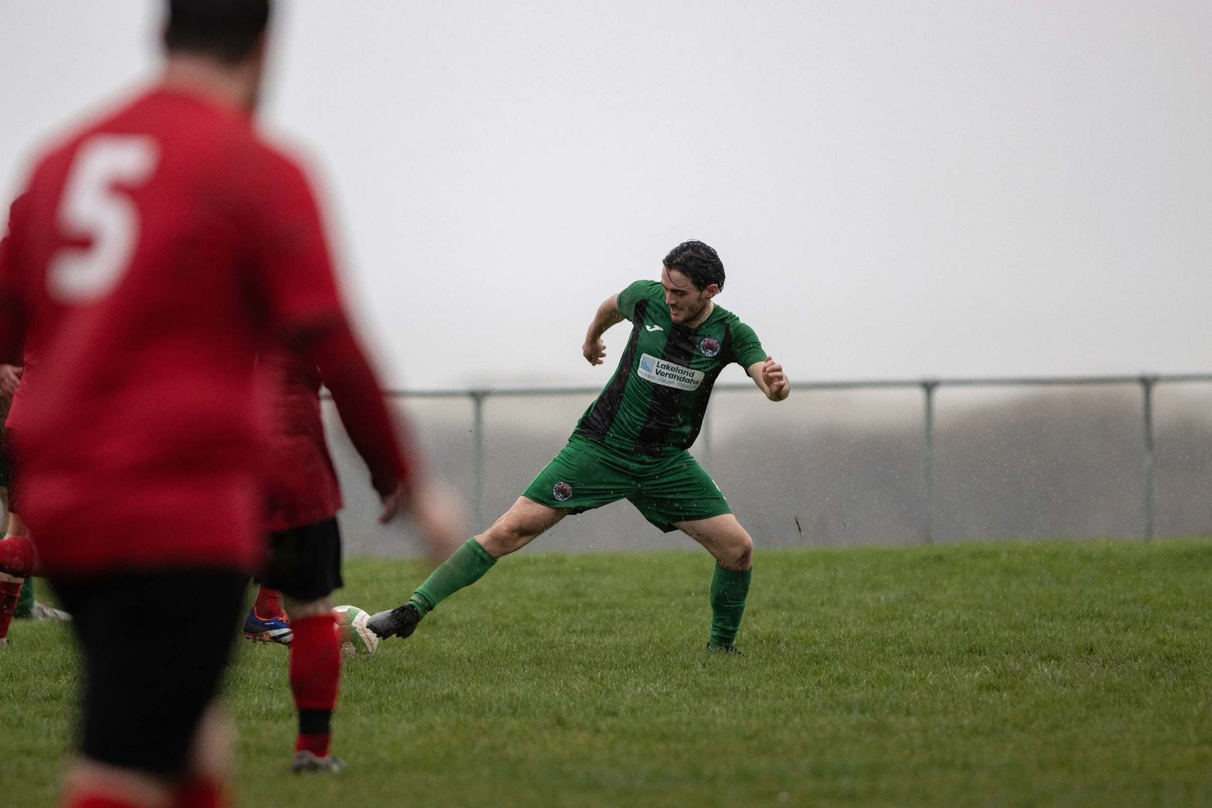 A soccer player in a green and black uniform is kicking a ball during a match on a grassy field on a rainy day. Other players are partially visible, with one in red in the foreground.