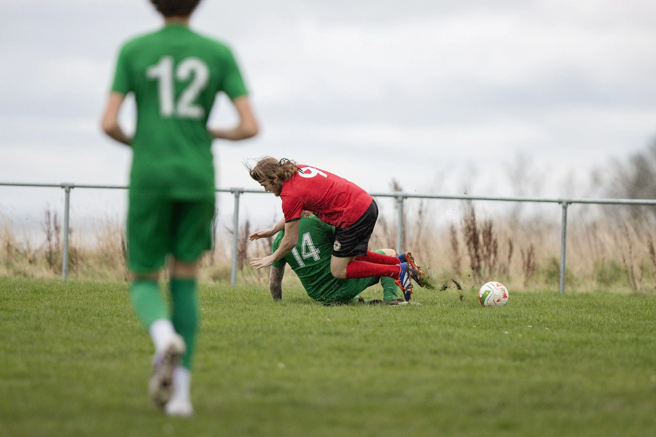 Two soccer players in green and red uniforms compete for the ball on a grassy field, with one player falling to the ground and the other in the foreground.