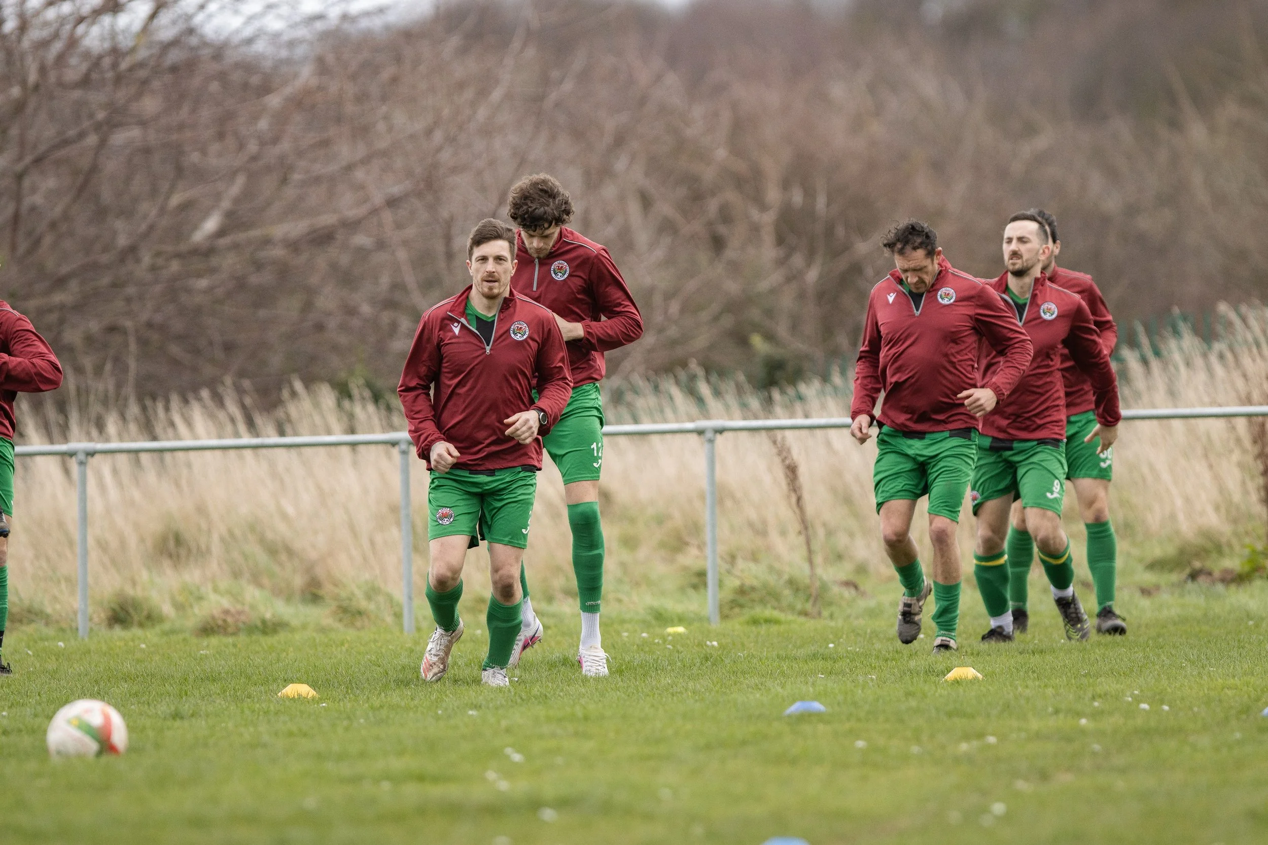 Soccer players in red and green uniforms jogging during practice on a grassy field. The background features leafless trees and a metal fence.