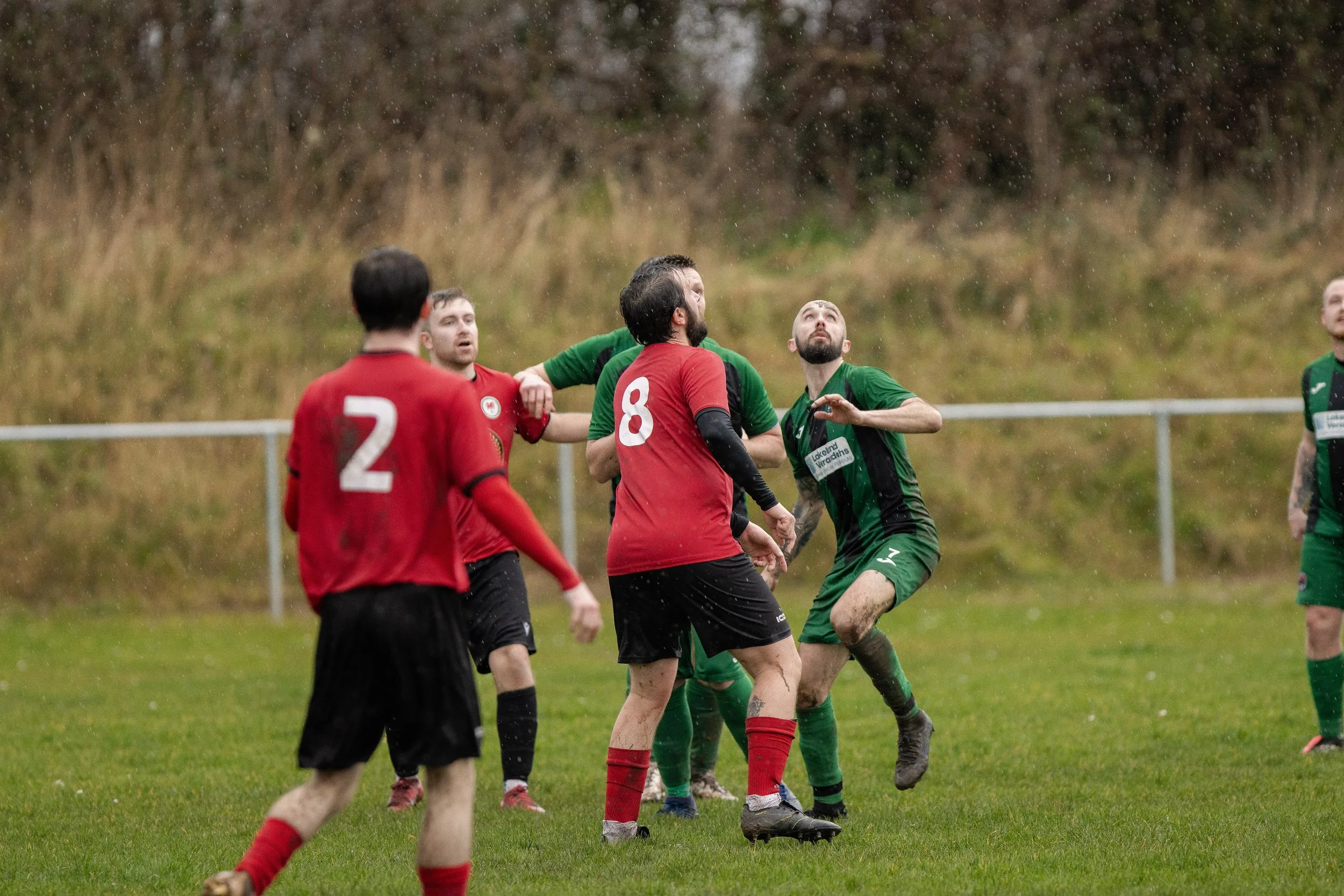 Soccer players in red and green jerseys contest a ball during a match on a rainy day, with some players looking upwards and wet grass in the background.