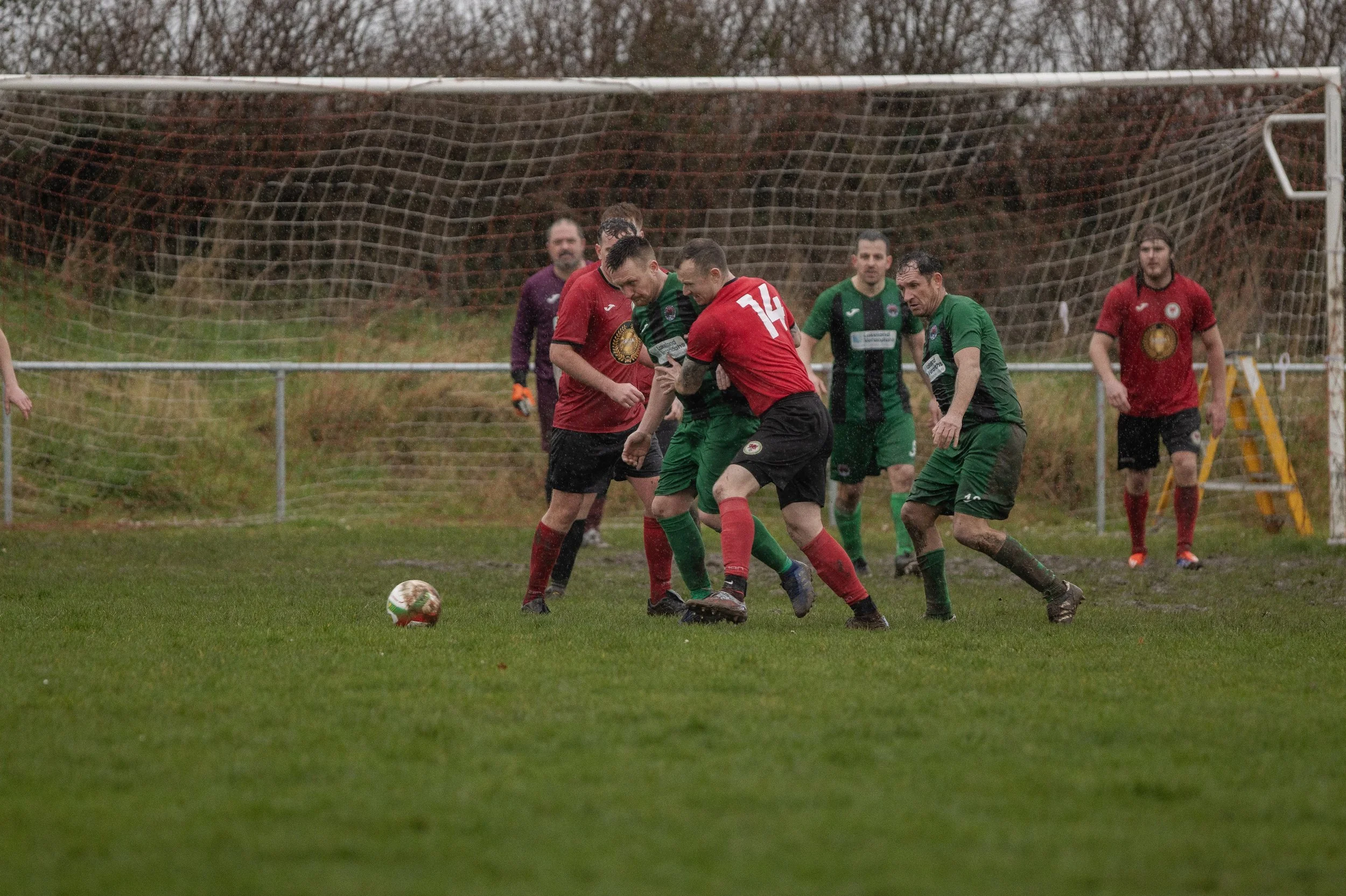 Soccer players in green and red jerseys competing for the ball on a muddy field near goalpost, with a goalkeeper in purple in the background.