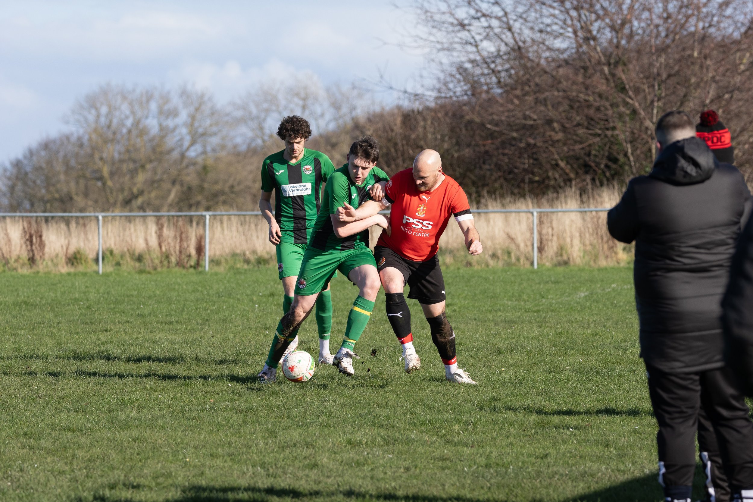 Two soccer players in green jerseys and black shorts compete for the ball while a referee in black watches nearby.