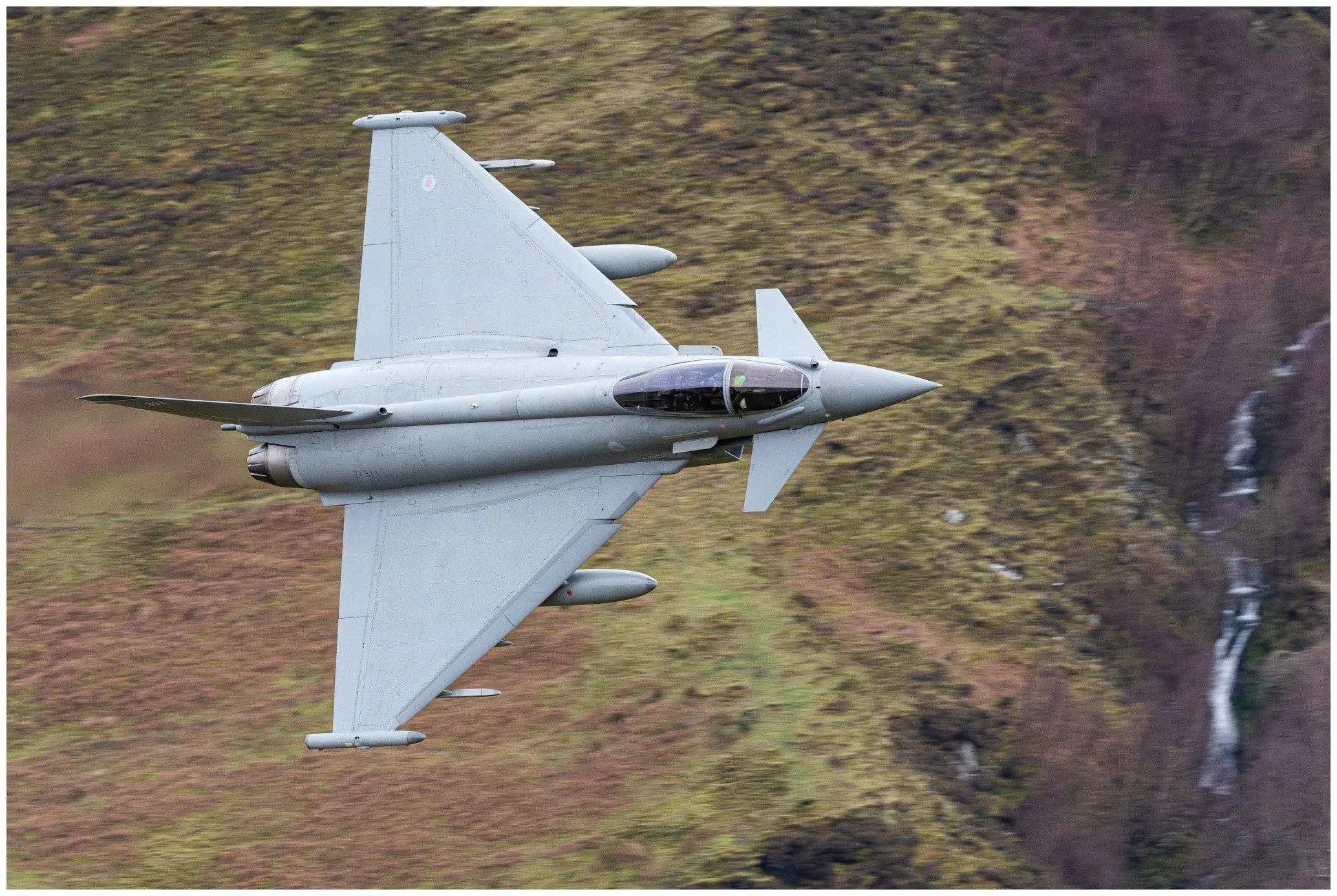 A gray fighter jet flying over a mountainous landscape with a waterfall on the side.