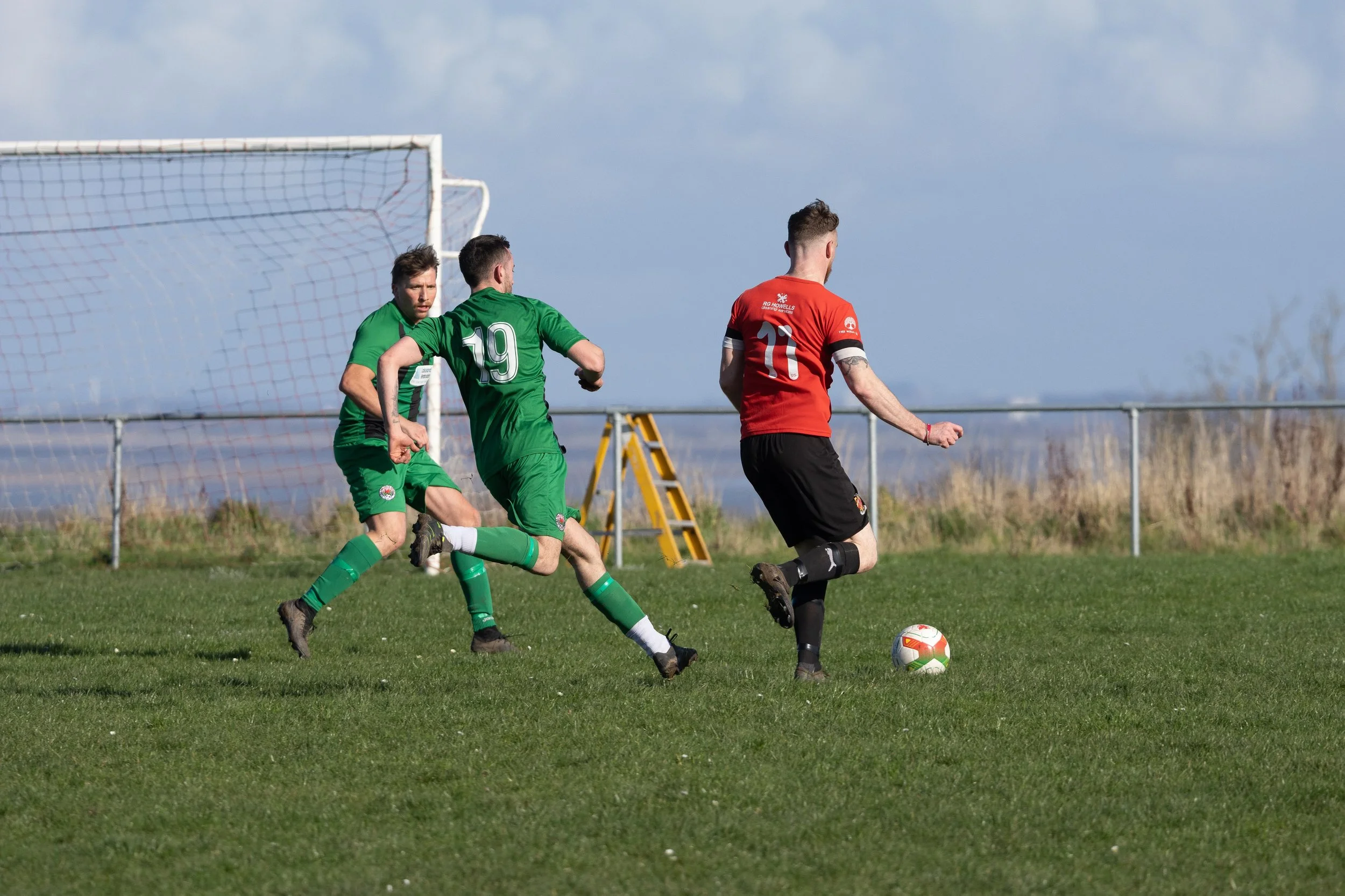 Three soccer players, two in green uniforms and one in red and black, are on a grassy field near the goal, with the player in red preparing to kick the ball while the other two players attempt to defend.