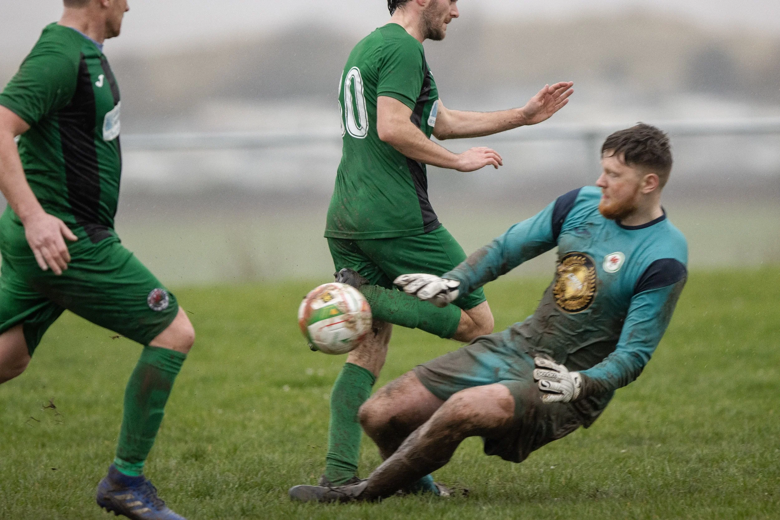 Soccer players in green and blue uniforms compete for the ball on a muddy field, with one player sliding to block the goalkeeper, who is on the ground.