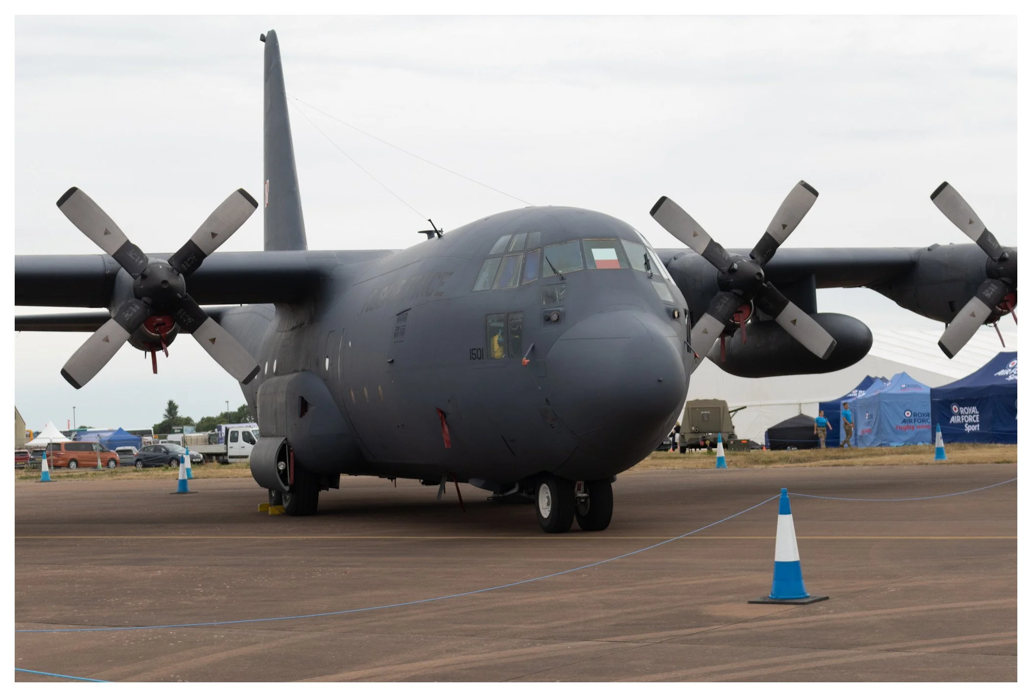 A large military cargo airplane parked on an airfield, with propellers and a dark gray body. There are tents and vehicles in the background.