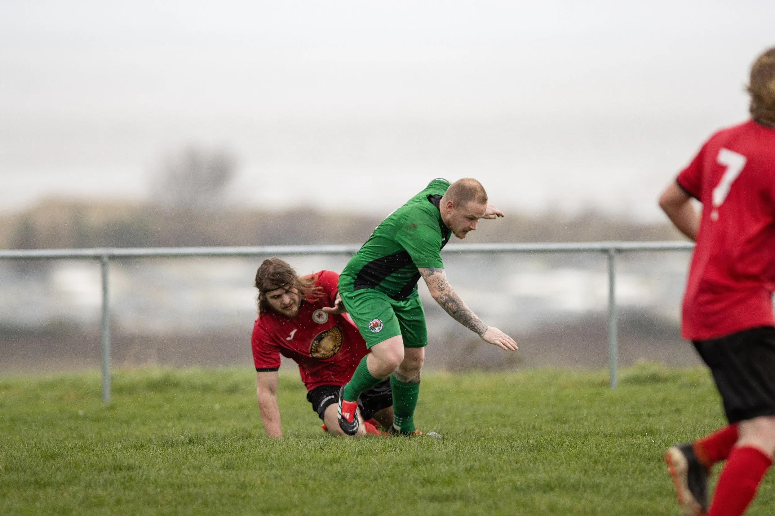 Two soccer players, one in a red jersey and the other in a green jersey, are competing on a grassy field. The player in red is on the ground, while the player in green is trying to maintain balance as he trips over the opponent. There is a blurred ba