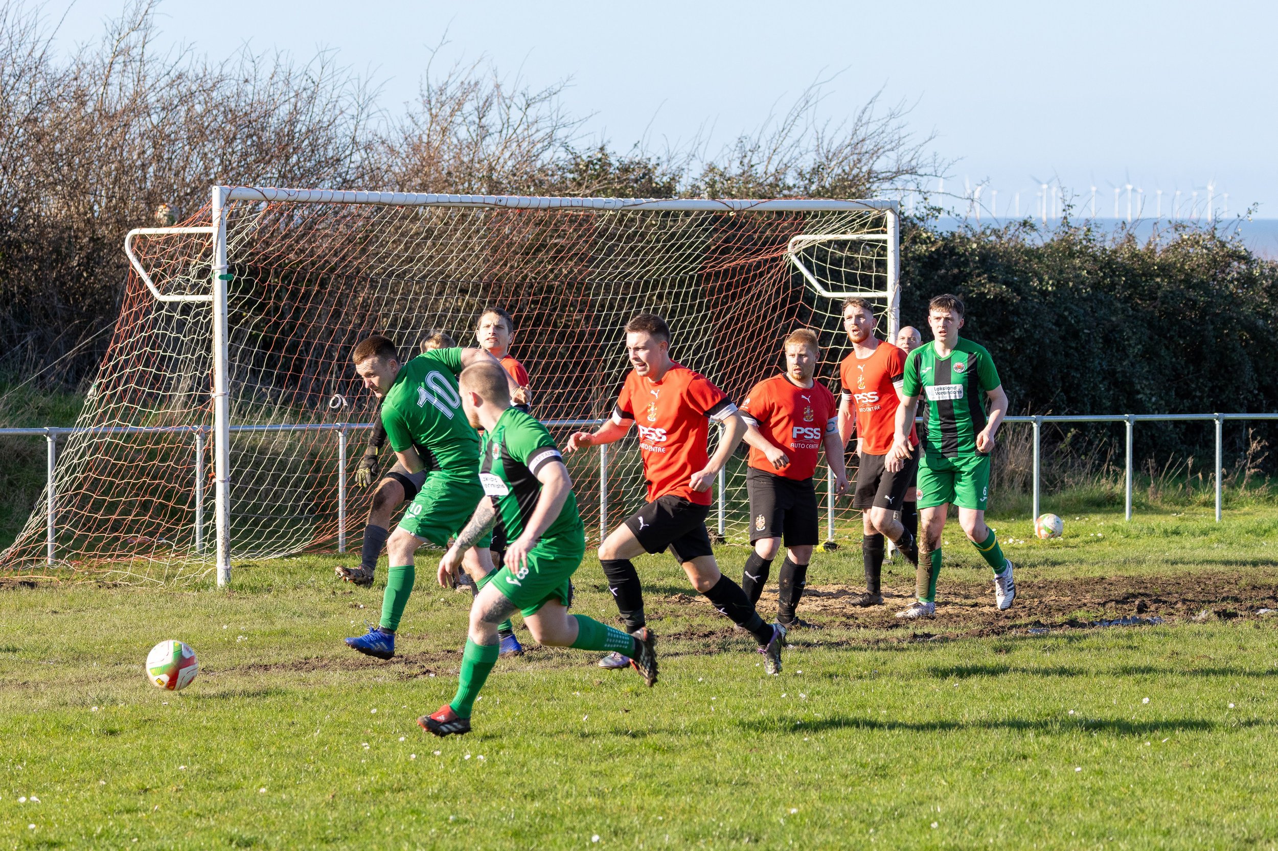 Soccer players in green and orange uniforms playing on a grassy field during a match, with a goal net and bushes in the background.