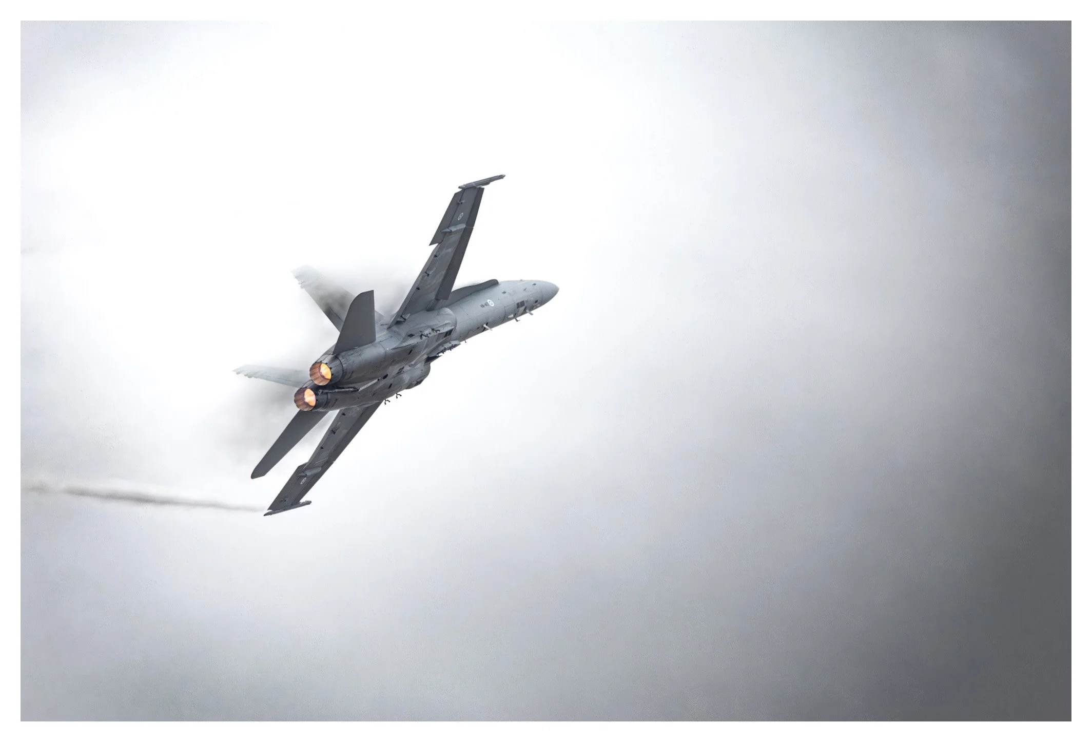 A military fighter jet flying through a cloudy sky, with visible engine exhaust and vapor trail.