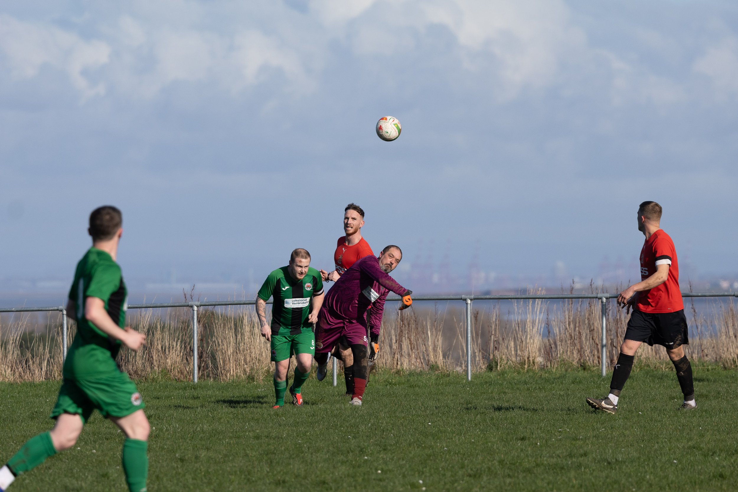 Five soccer players on a grassy field, with a metal fence and tall grass behind them, playing during the day with cloudy sky. One player in green is running, another in green is near the goal, a player in red, another in red, and a goalkeeper in purp