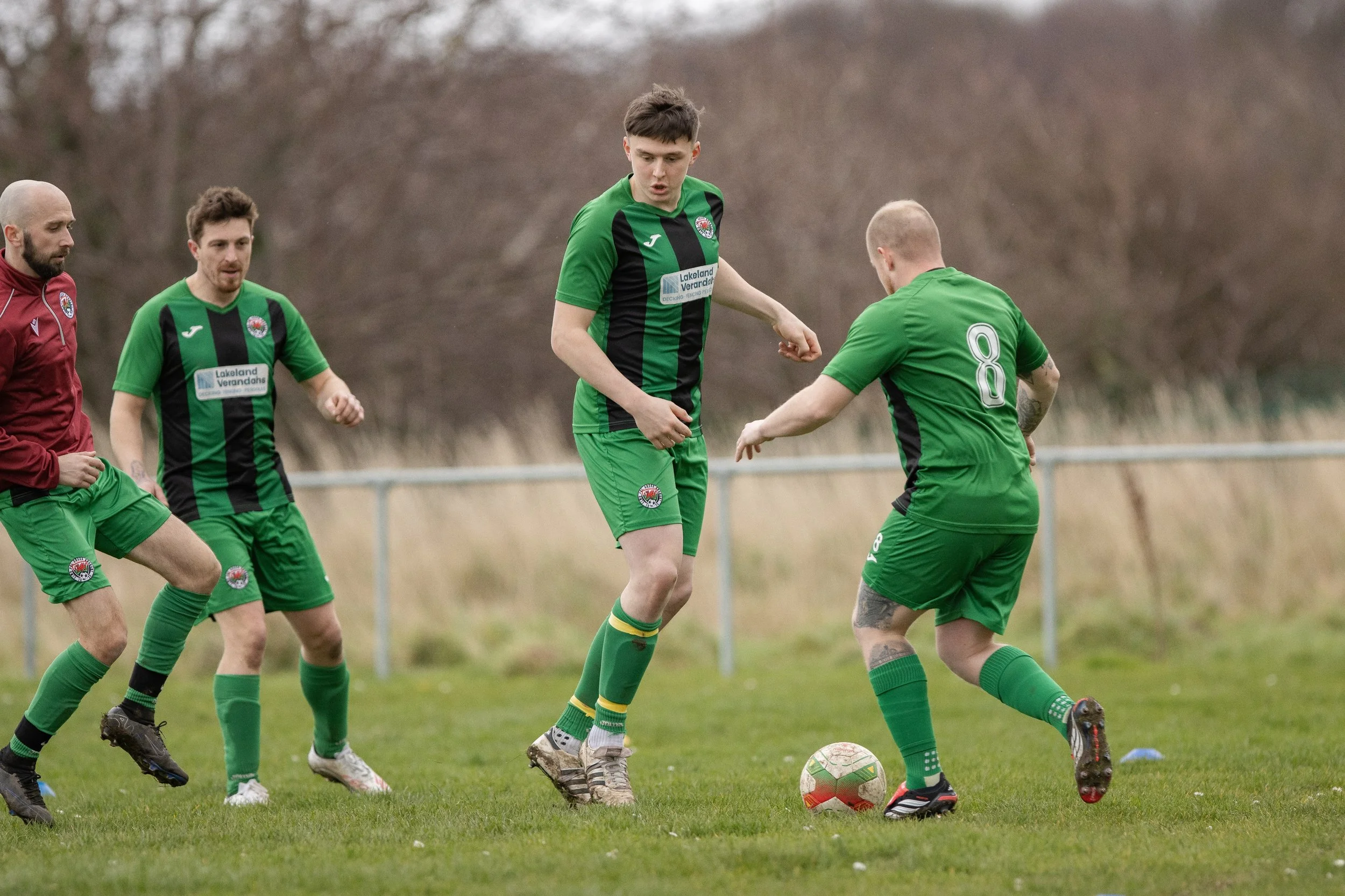 Four soccer players in green uniforms playing on a grassy field, with one player dribbling the ball while the others look on.