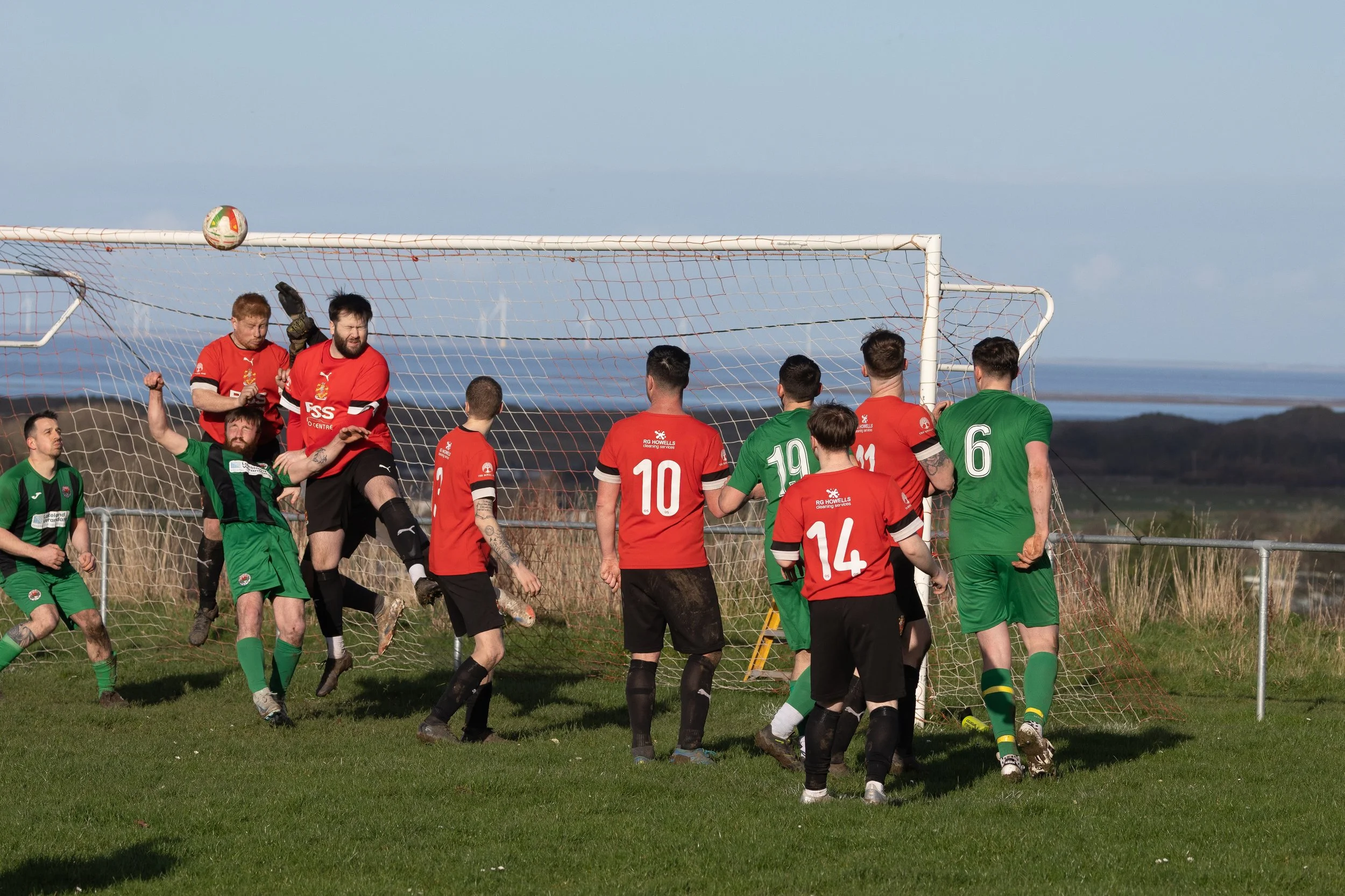 Soccer players in red and green uniforms contest a ball in front of the goal outdoors on a grassy field with a scenic landscape and wind turbines in the background.