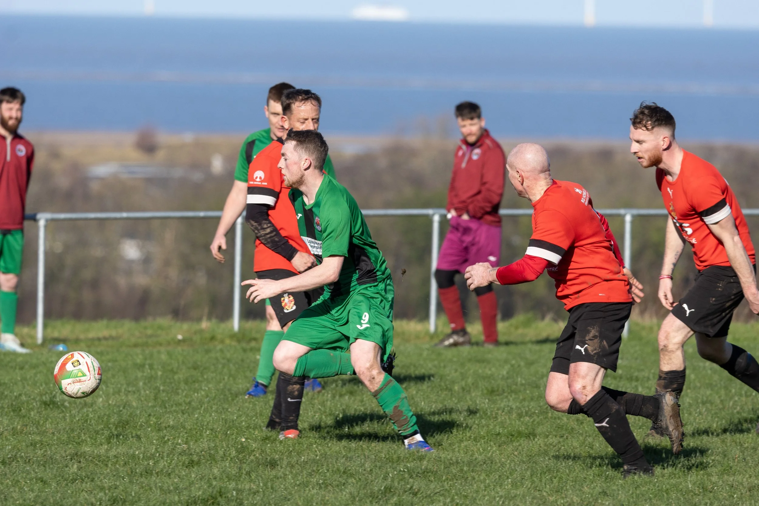Soccer players in red and green jerseys competing for the ball on a grassy field during a daytime match.