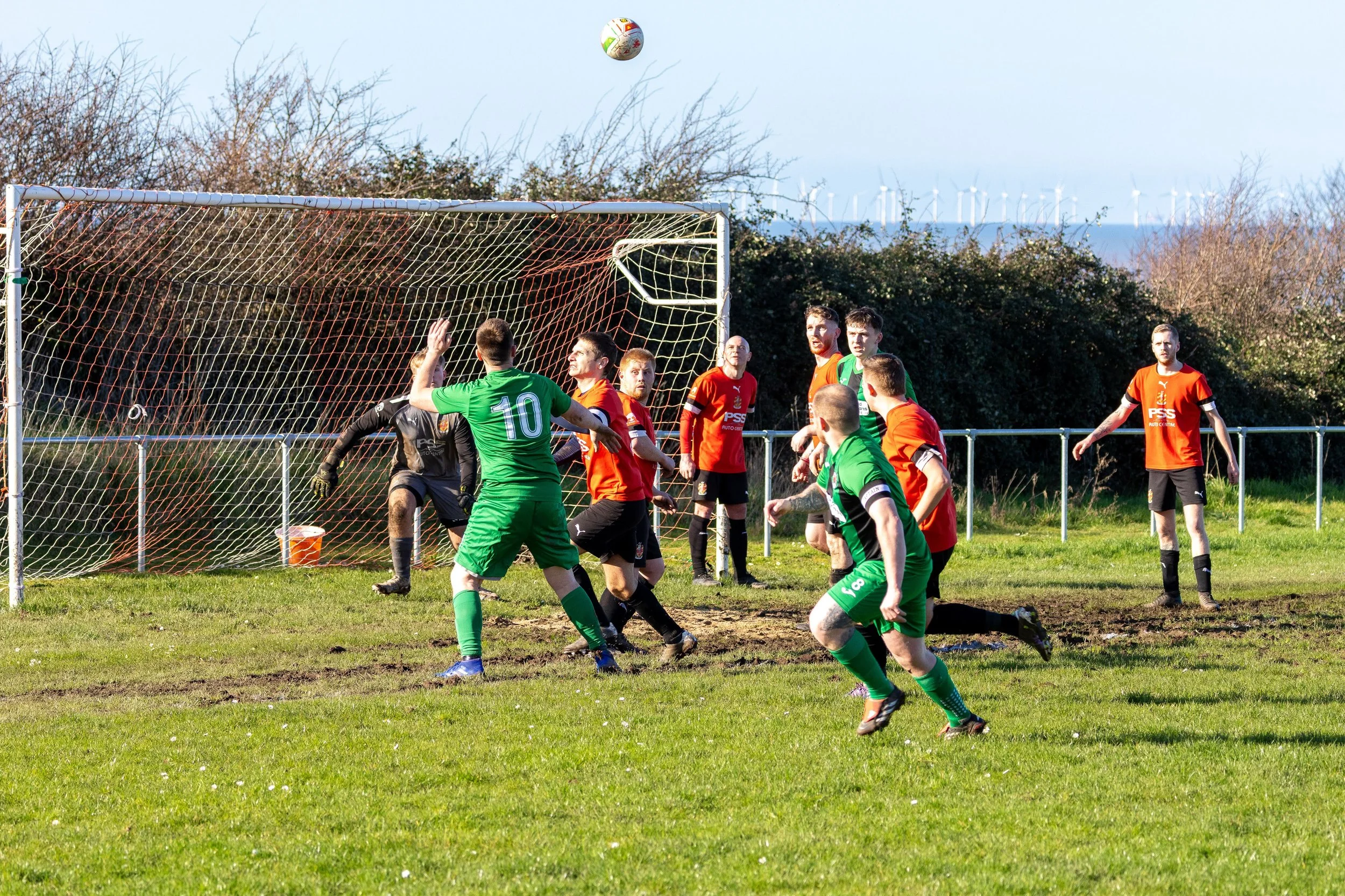 Soccer players competing for ball near the goal post on a grassy field during daytime, with some players in green and others in orange and red jerseys, and wind turbines visible in the background.
