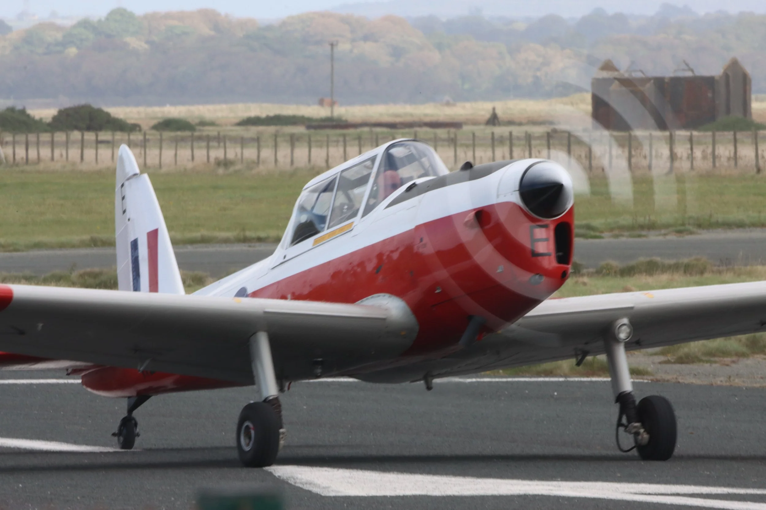A vintage aircraft on a runway with a person inside, propeller spinning, and a rural landscape with fields and hills in the background.