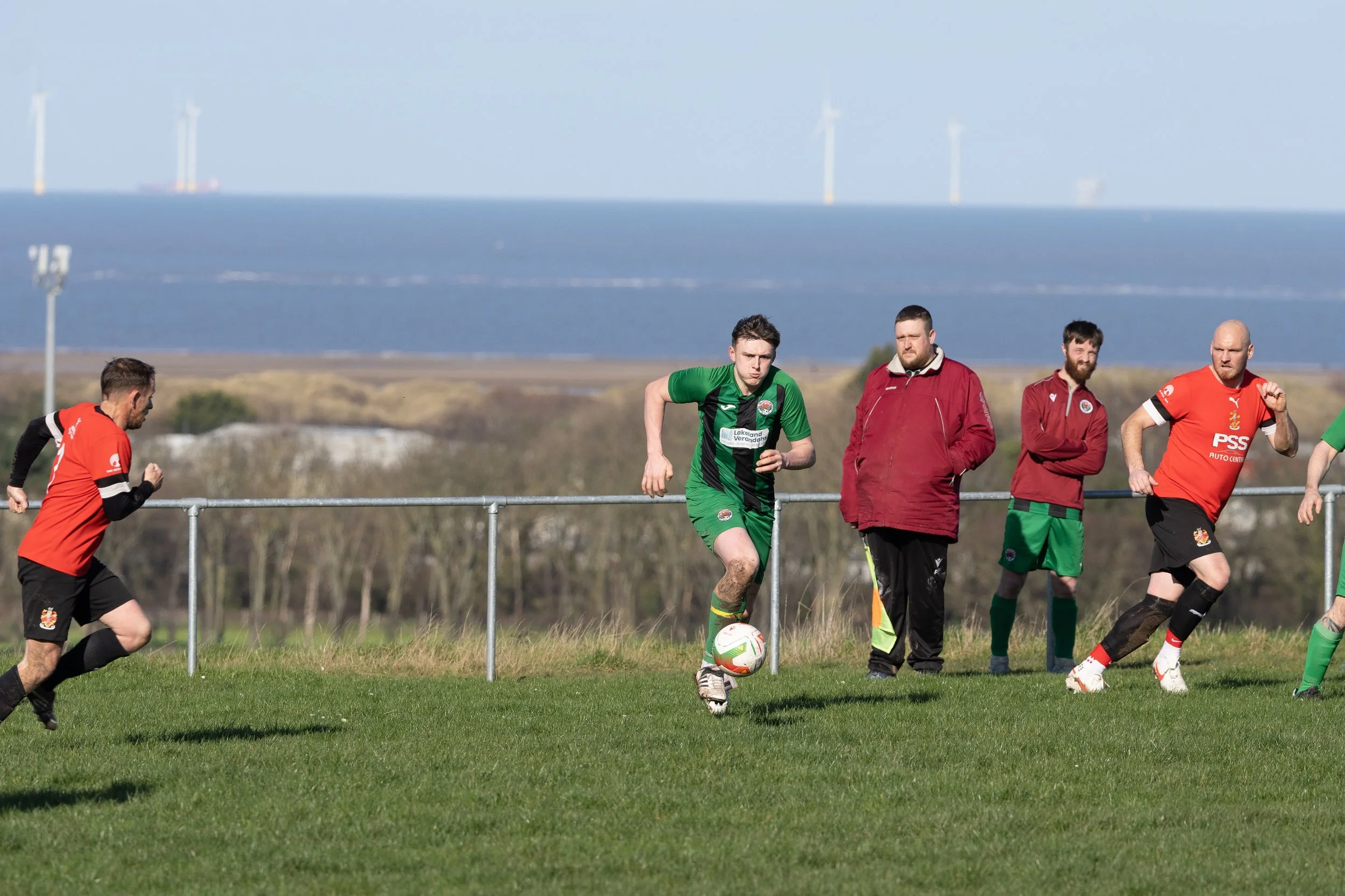 Soccer players practicing on a grass field with a fence, water, and wind turbines in the background. One player in a green uniform controls the ball, while others in red and green uniforms watch or run.