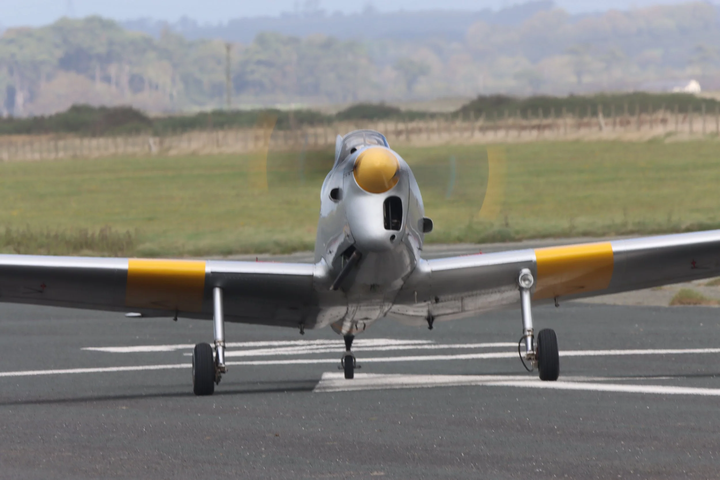 A vintage fighter jet aircraft on an airstrip, facing directly toward the camera with spinning propellers, yellow mask, and landing gear visible.
