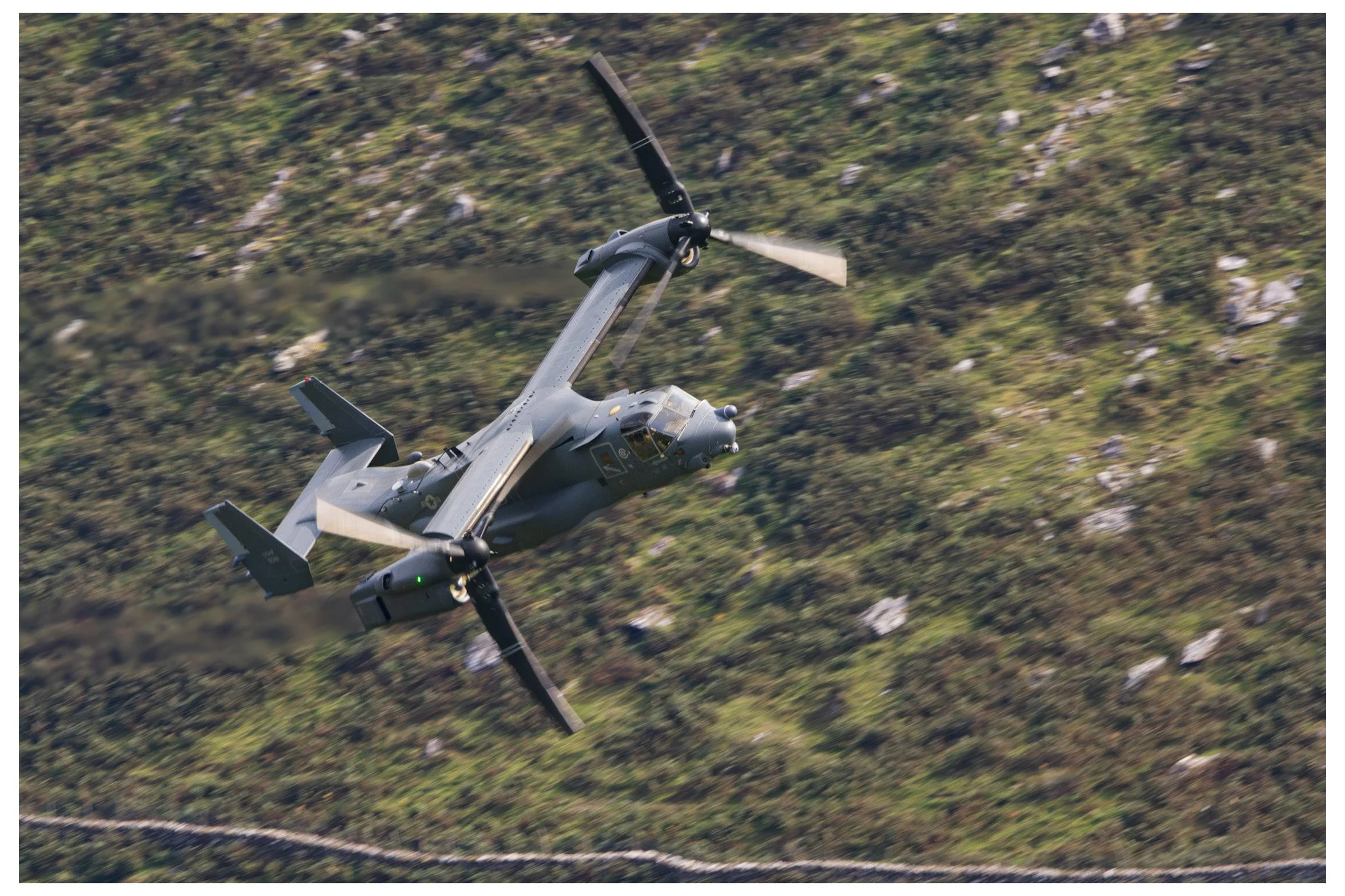 A military tiltrotor aircraft flying over a mountainous landscape with green vegetation and rocks.
