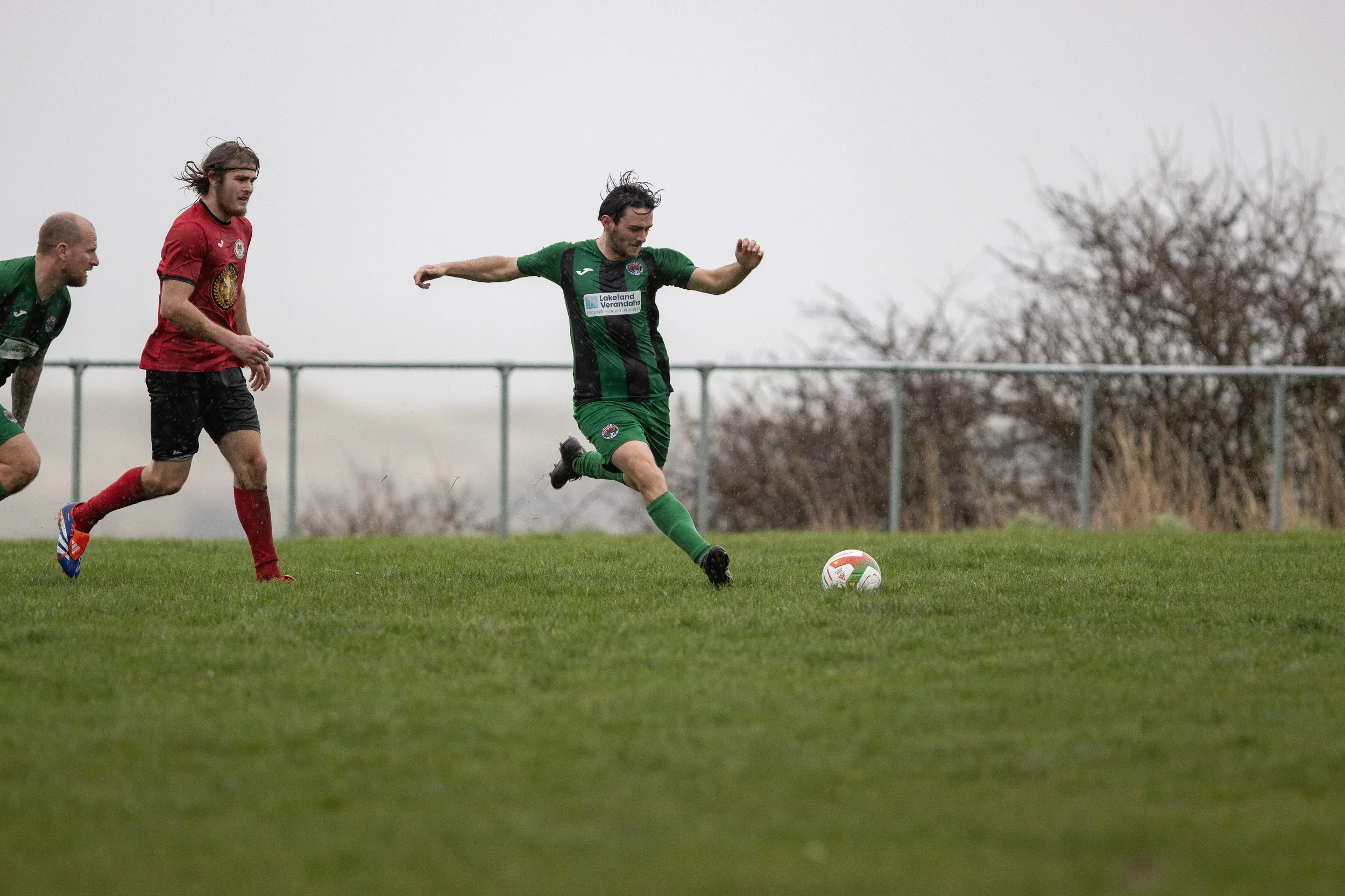 Soccer players in action on a grassy field during a match, with one player in a green and black uniform kicking the ball, and two players in red and black uniforms pursuing.
