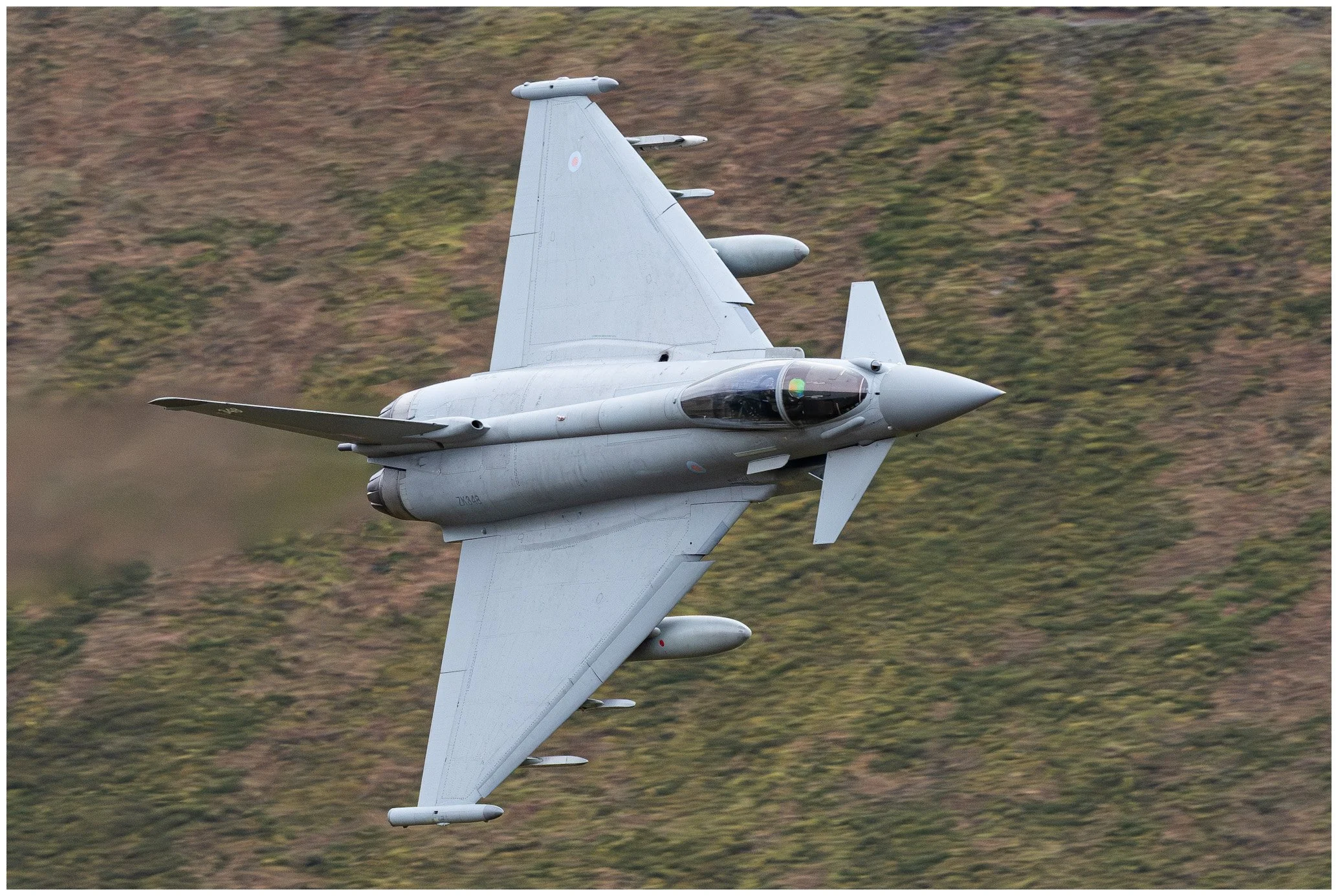 A gray military fighter jet flying at an angle with a green and brown landscape blurred in the background.