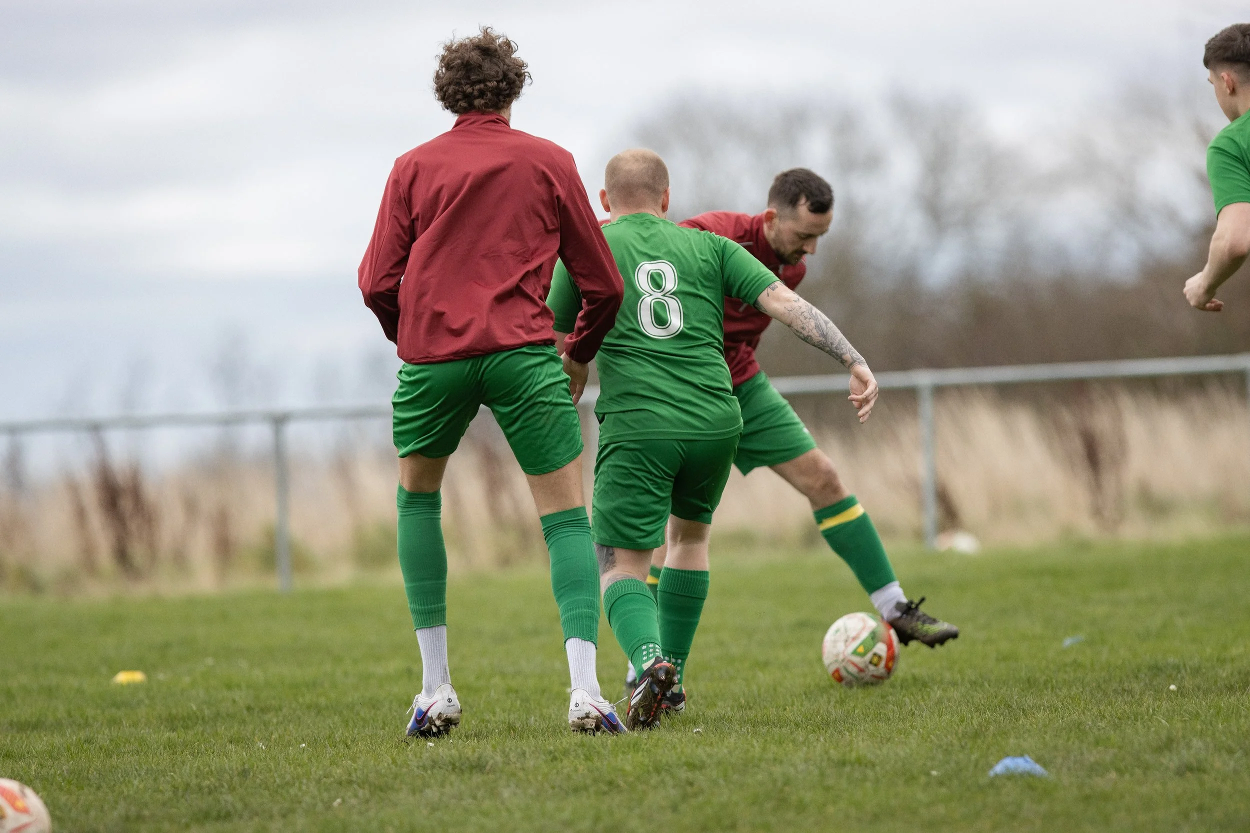 Soccer practice with three players on a grass field, one wearing a green jersey with the number 8, and two others in red jackets, engaging in a drill.
