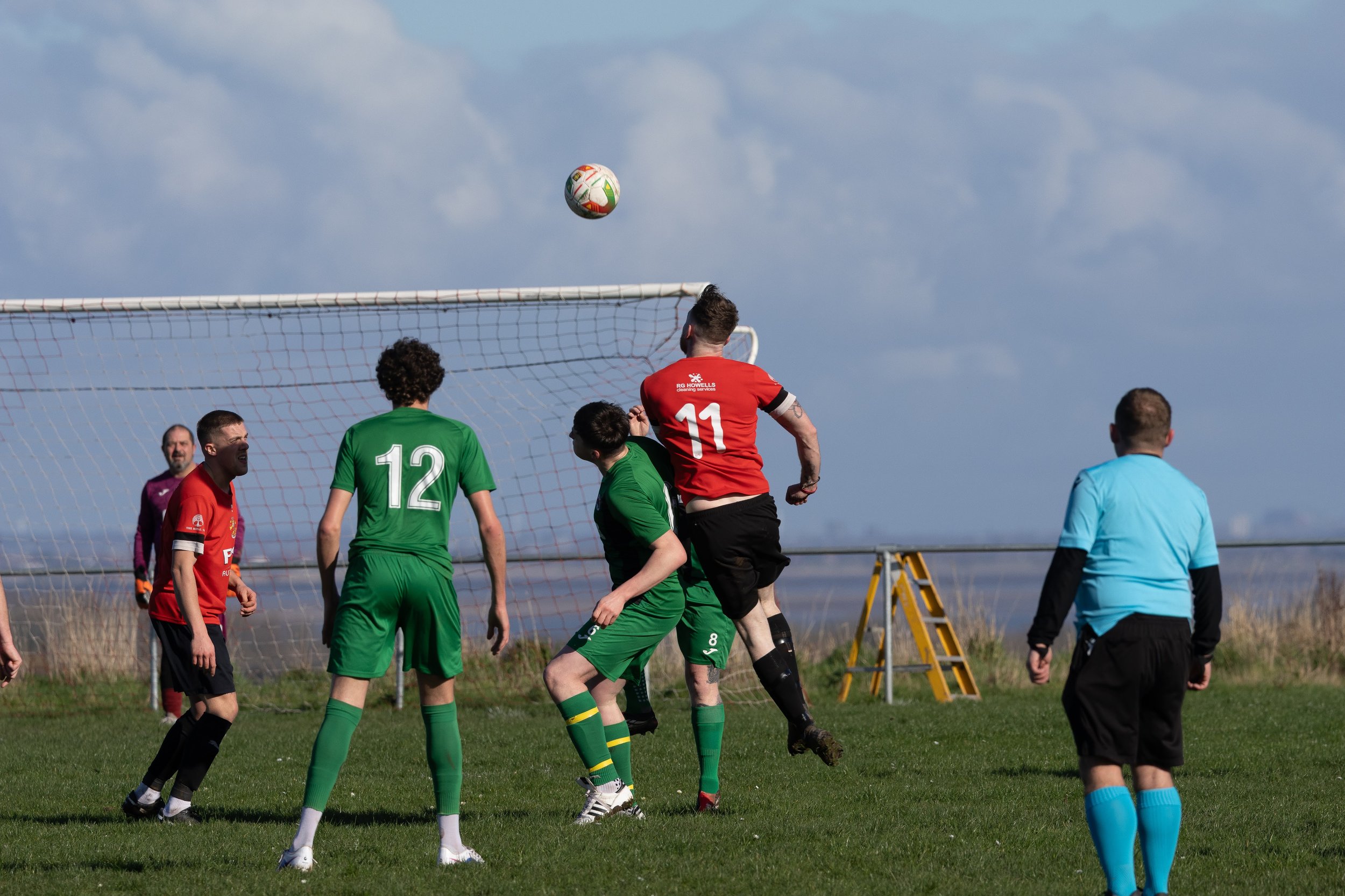Soccer players in green and red jerseys competing for the ball during a game on a grassy field, with a goal and a clear sky in the background.