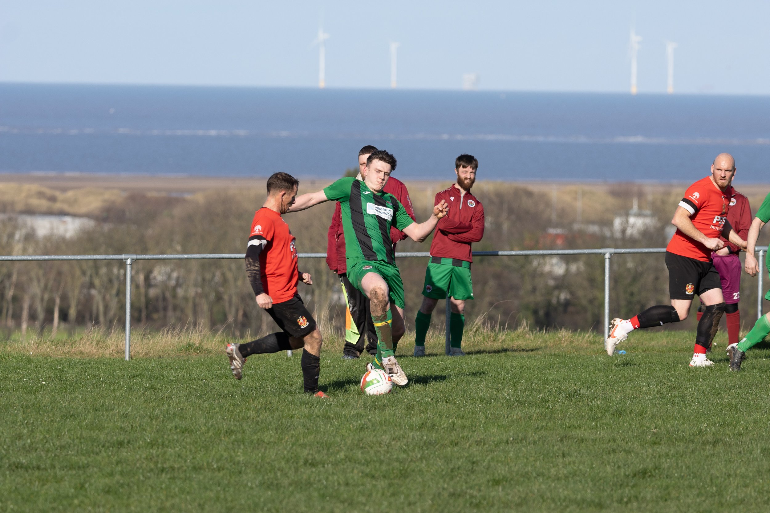 Soccer players on the field during a match, with a coastline and wind turbines in the background.