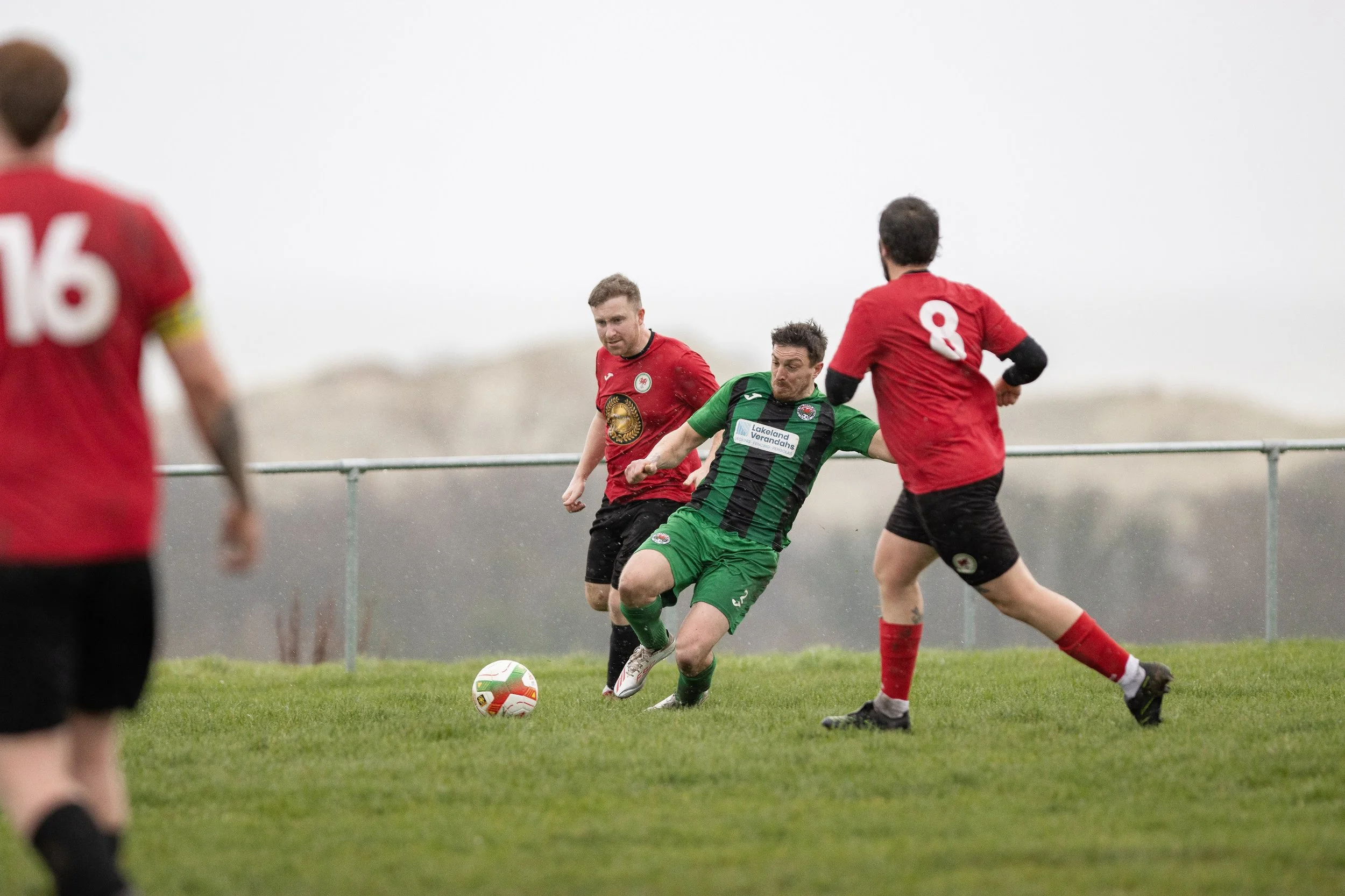 Soccer players in red and green jerseys competing for the ball on a grassy field outdoors.