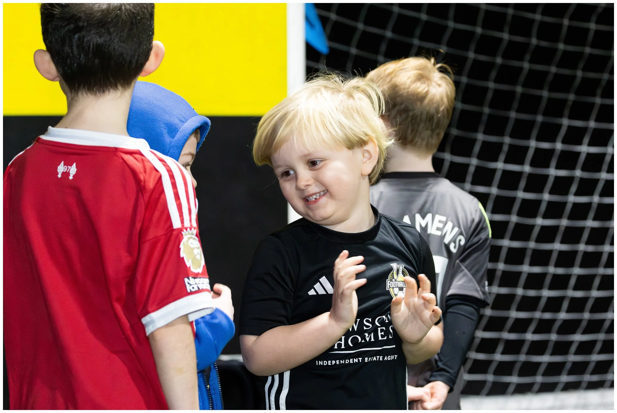 Children in sports attire standing in front of a soccer goal, engaging in a conversation at an indoor sports facility.