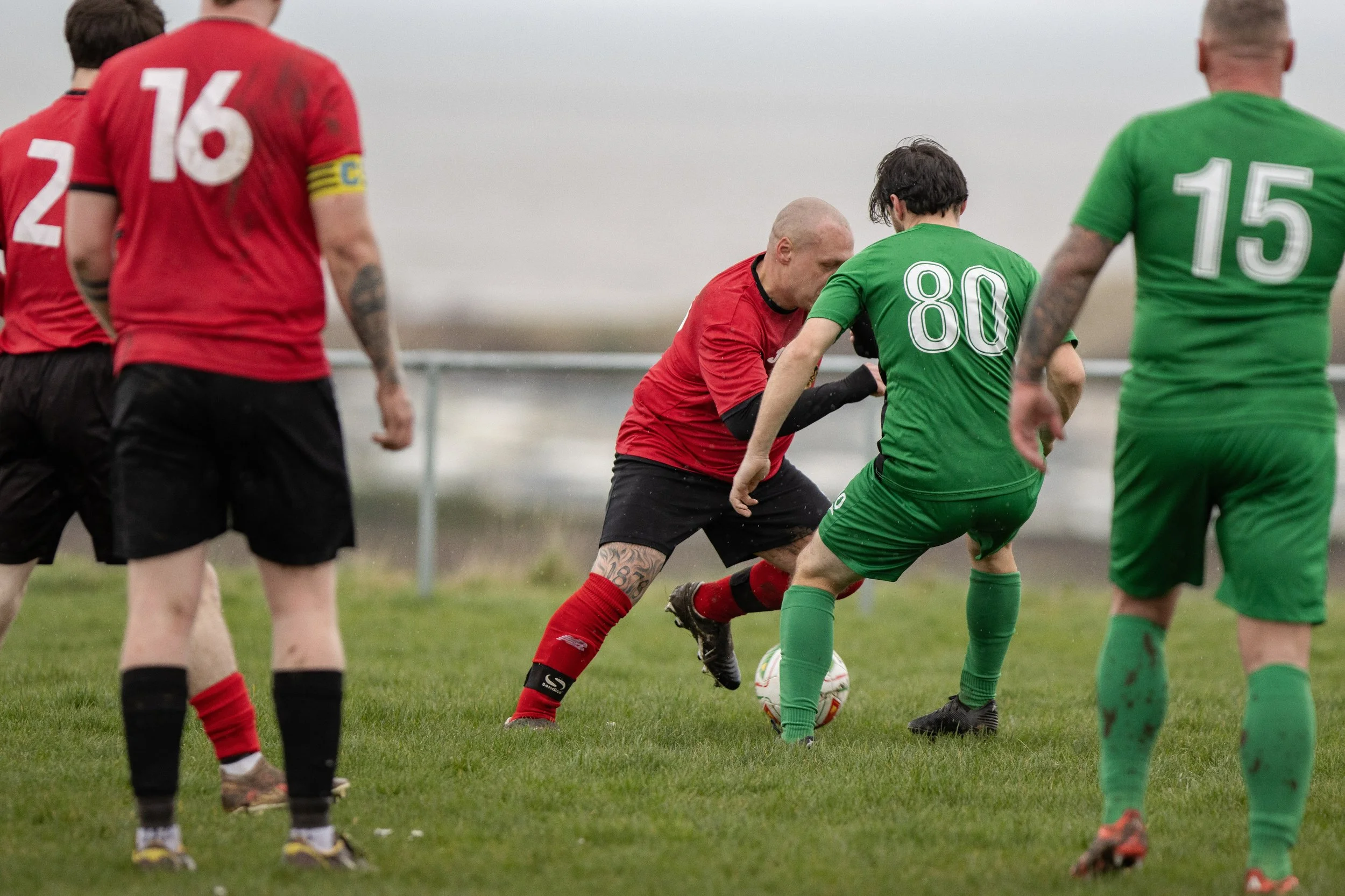 Soccer players competing for ball on field, some with mud-stained uniforms, overcast sky.