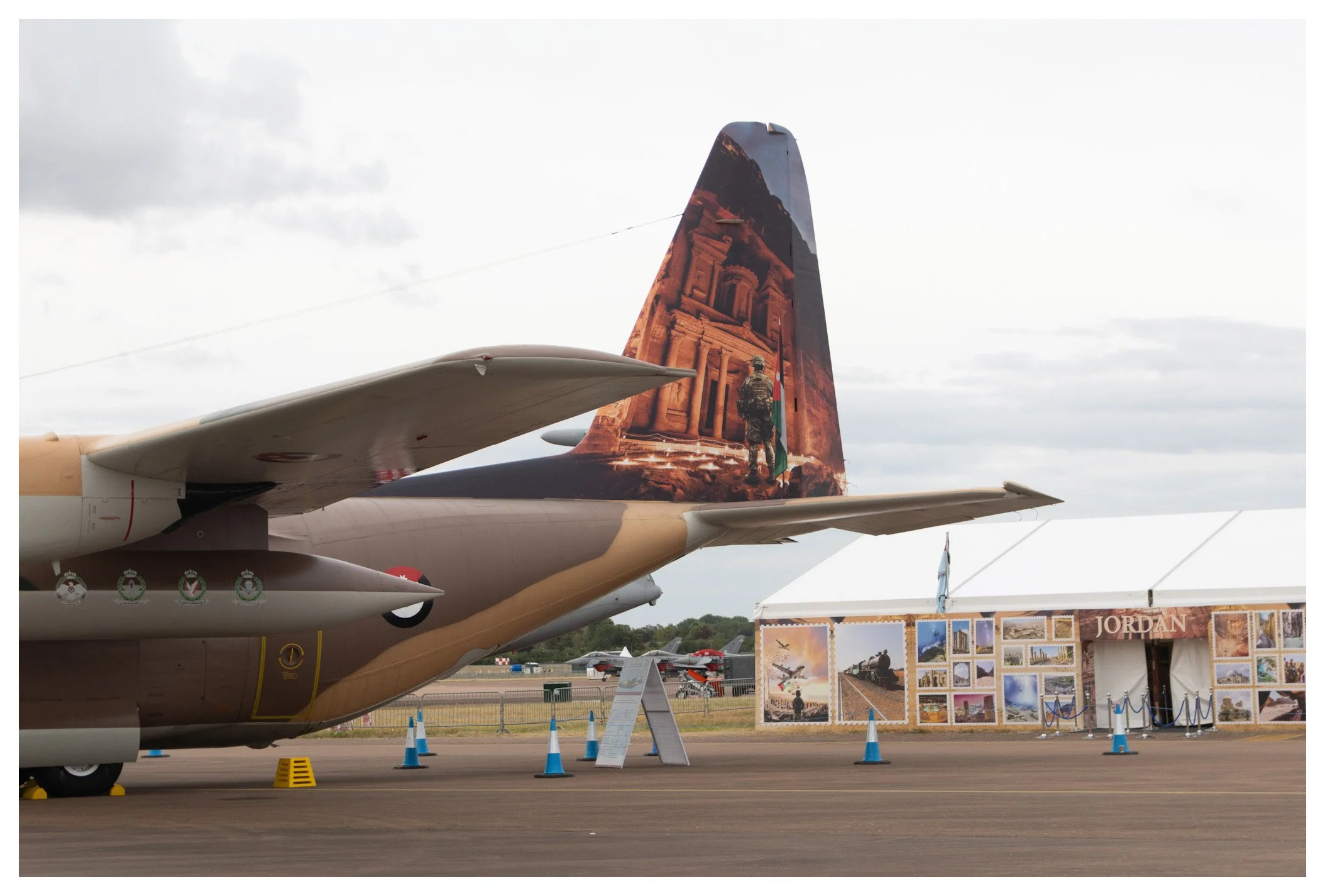 A military aircraft on display at an airshow, with a sculpture of a soldier and ancient Jordanian architecture painted on its tail, and a tent with images of Jordanian landmarks in the background.