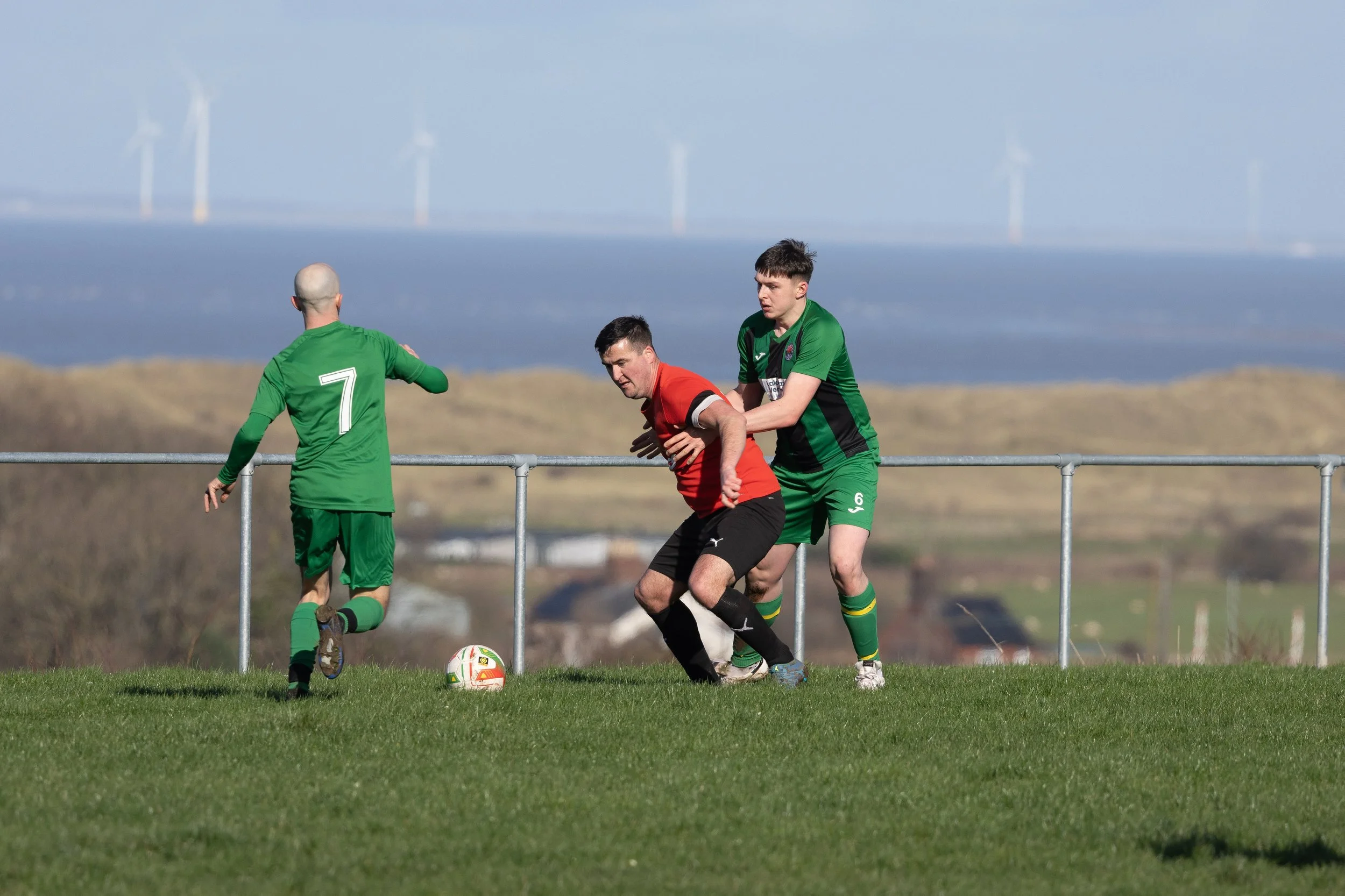 Soccer players competing for the ball during a match on a grassy field with a rural landscape and wind turbines in the background.