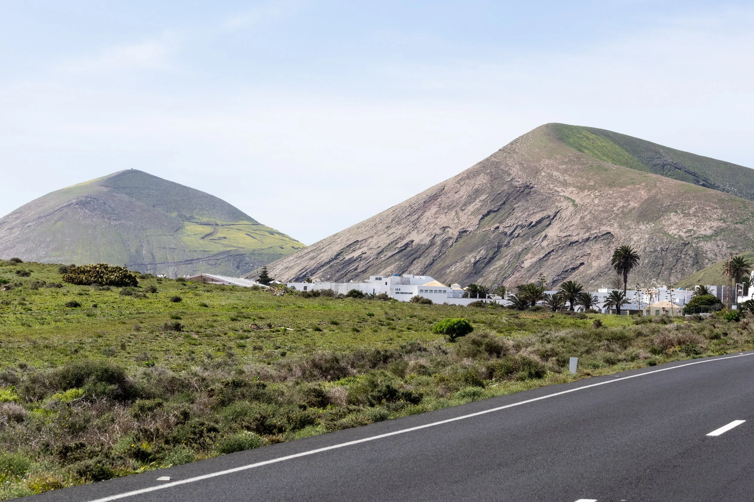 Landscape with mountains, green vegetation, white buildings, and a curved road in the foreground.