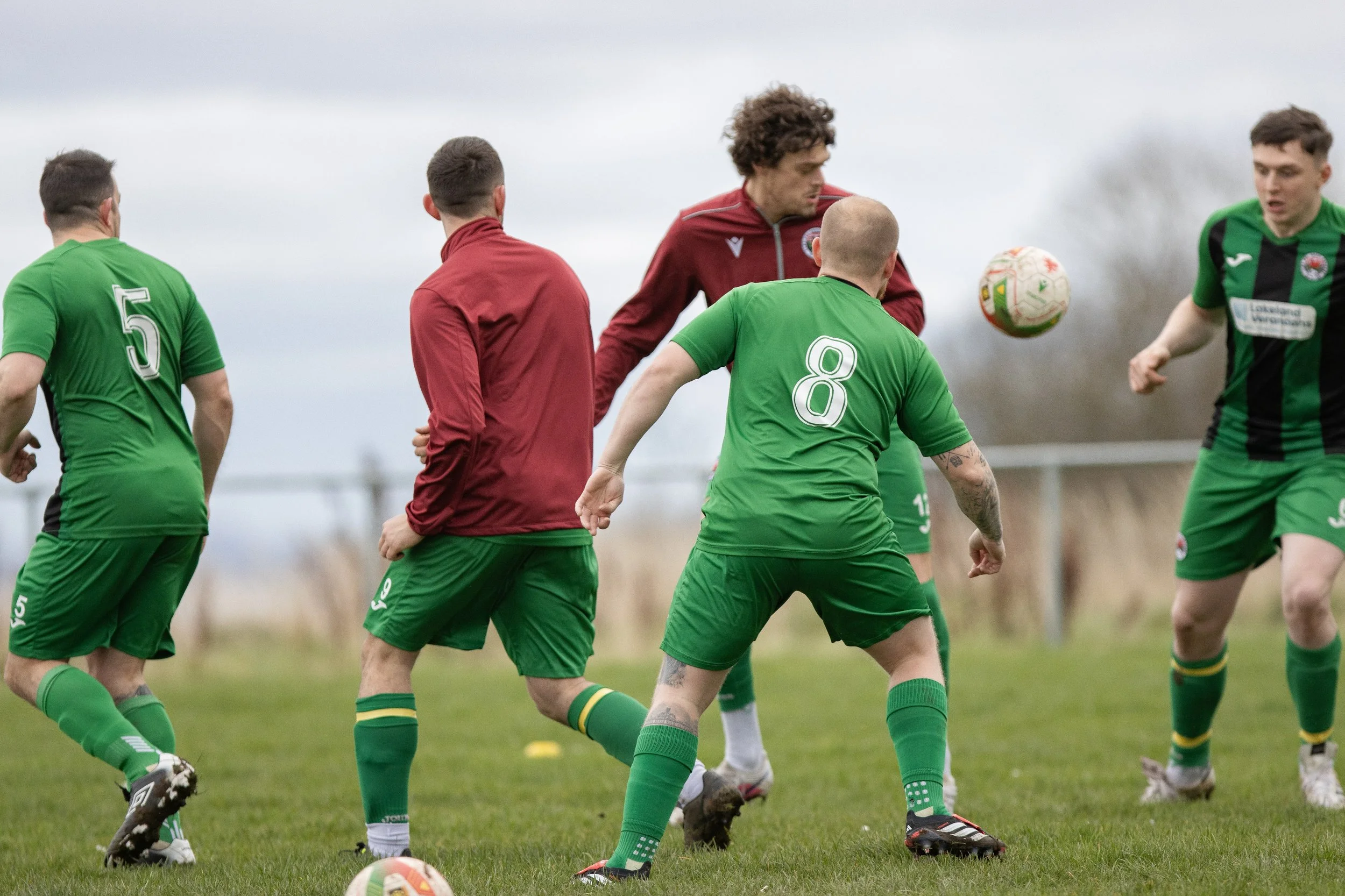 Soccer players in green and red jerseys competing for ball during practice or game on a grassy field under cloudy sky.