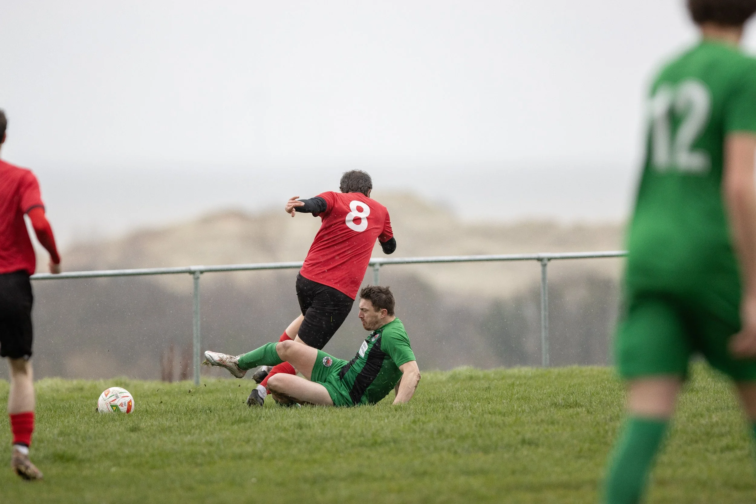 A soccer match with players wearing red and green uniforms, with one player in red appearing to fall or be tackled by a player in green who is seated on the ground, and other players nearby on a grassy field under an overcast sky.