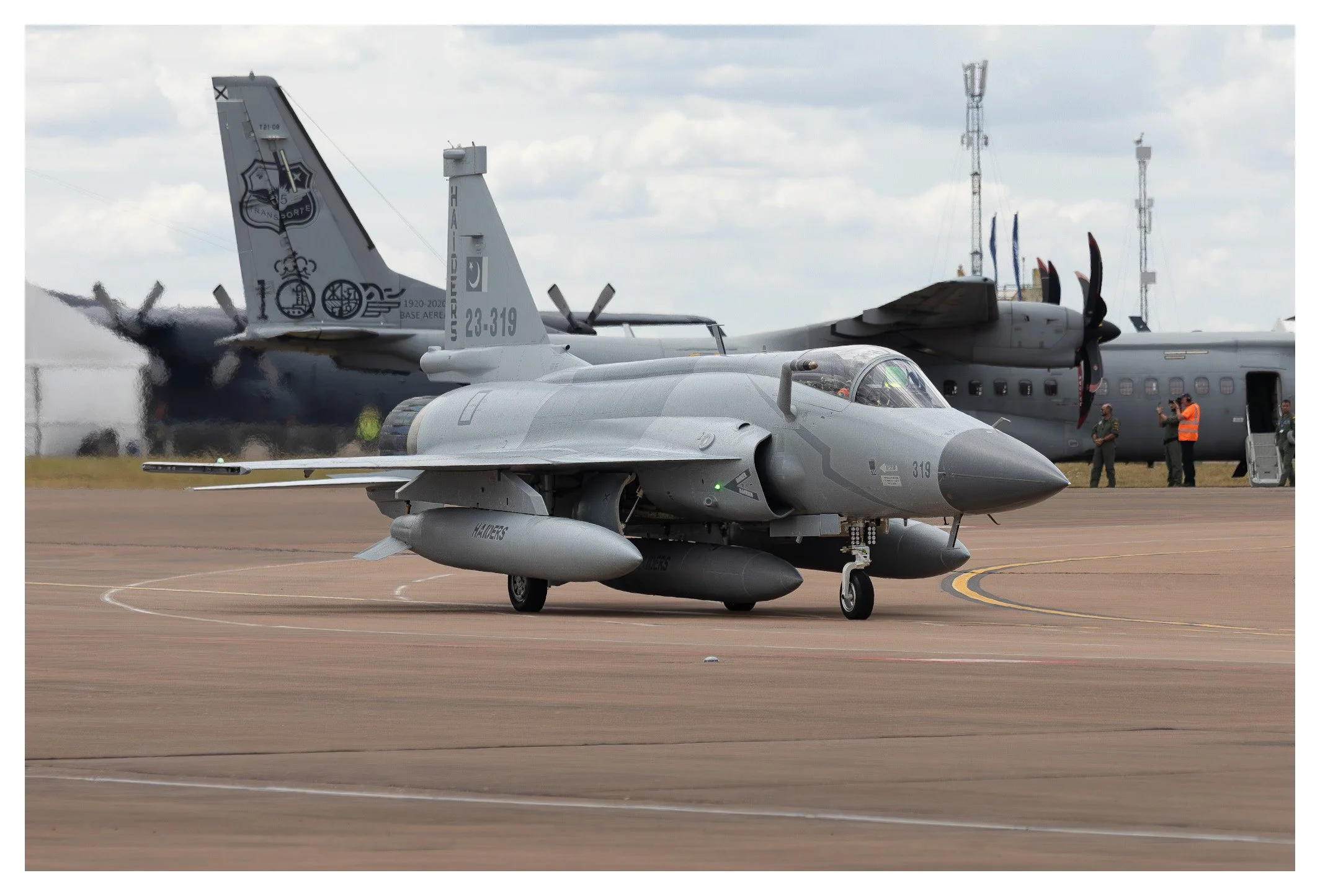 A military fighter jet taxiing on an airstrip with a larger aircraft in the background.
