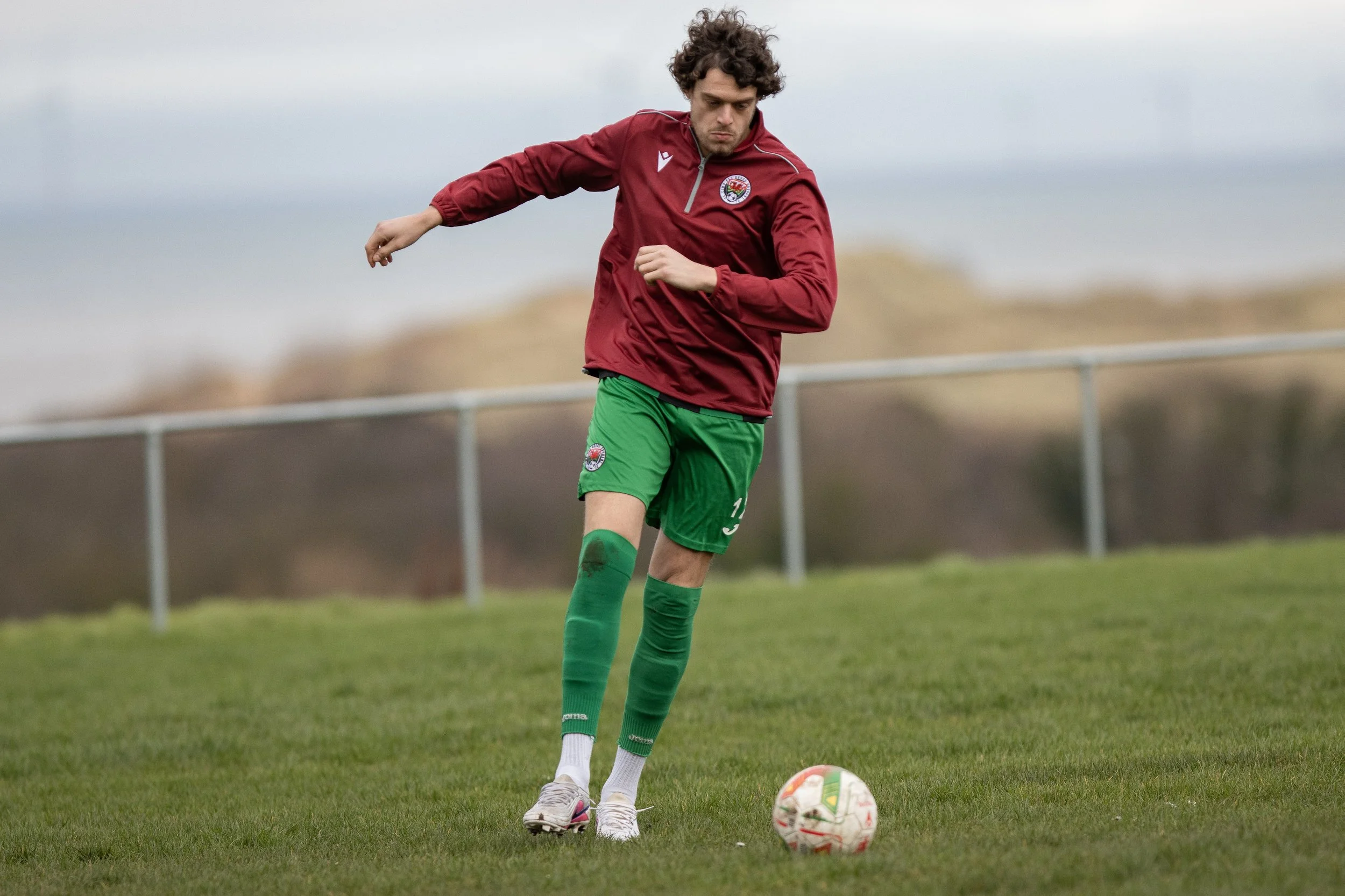 A man in a red sports jacket and green shorts is playing soccer on a grassy field, preparing to kick a white and red soccer ball.